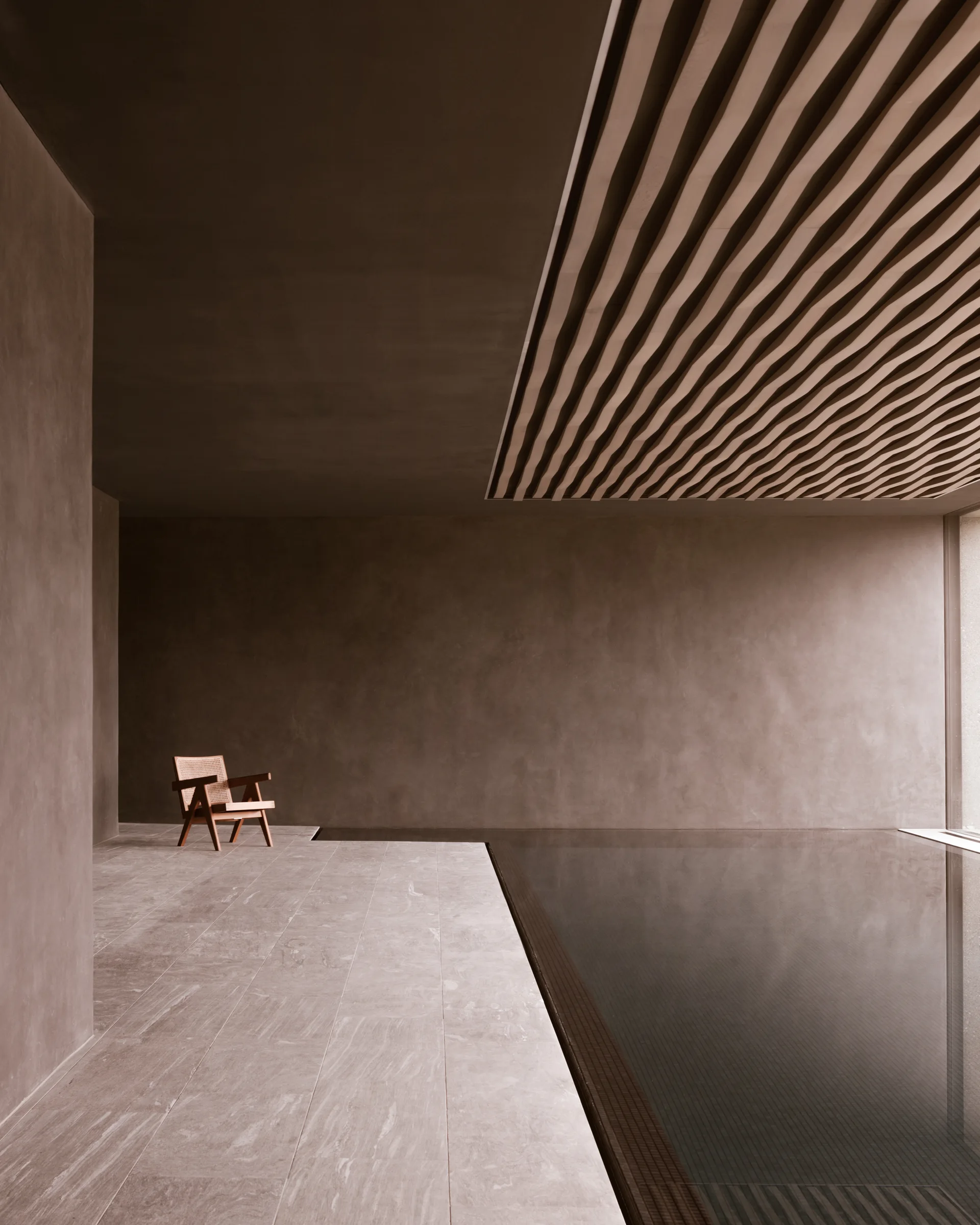 A minimalist indoor pool area with a wooden lounge chair on stone tiles next to still water under a slatted ceiling.
