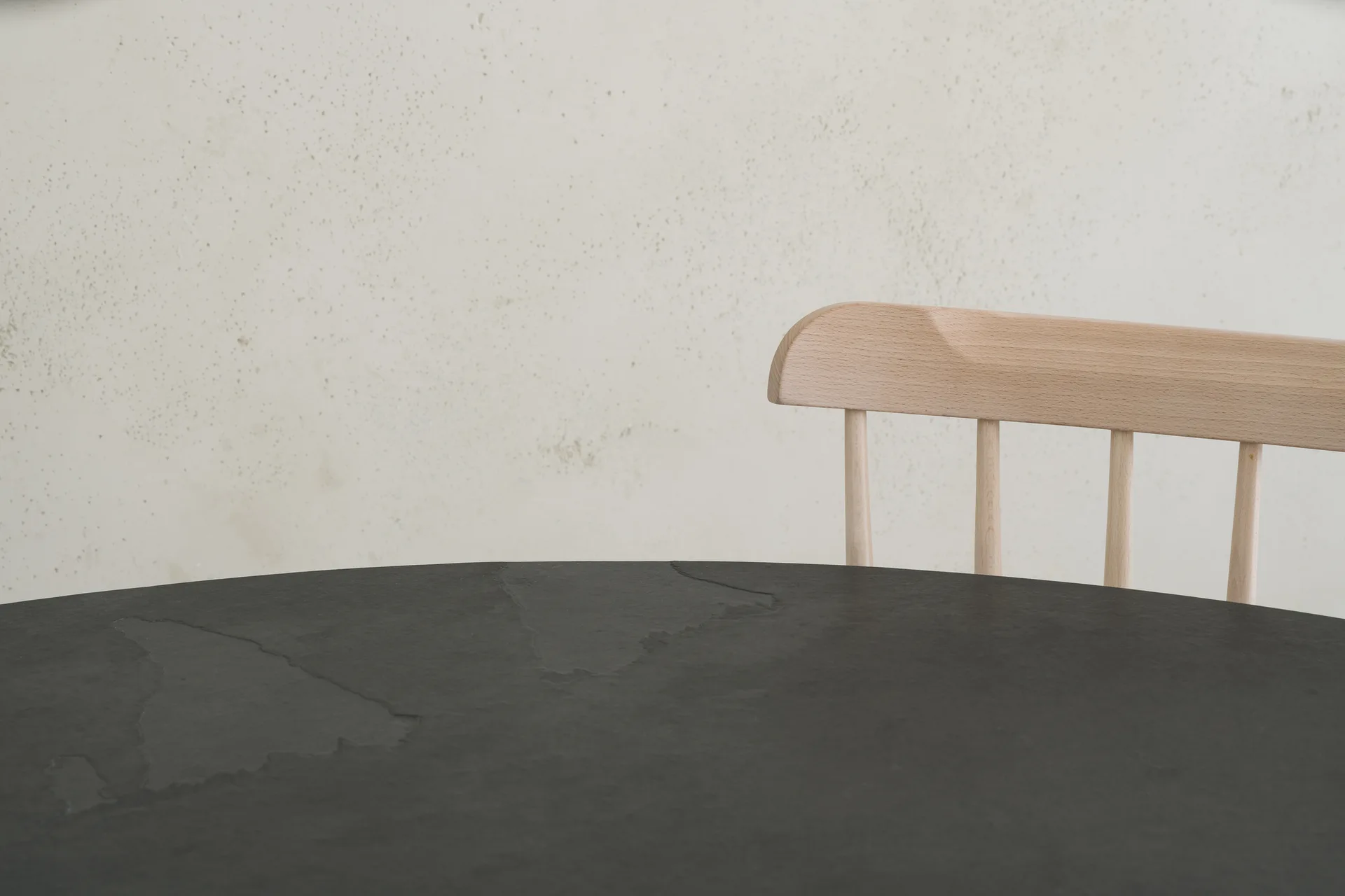 A close-up of a matte charcoal stone dining table paired with a light natural wood spindle-back chair against a textured off-white plaster wall.