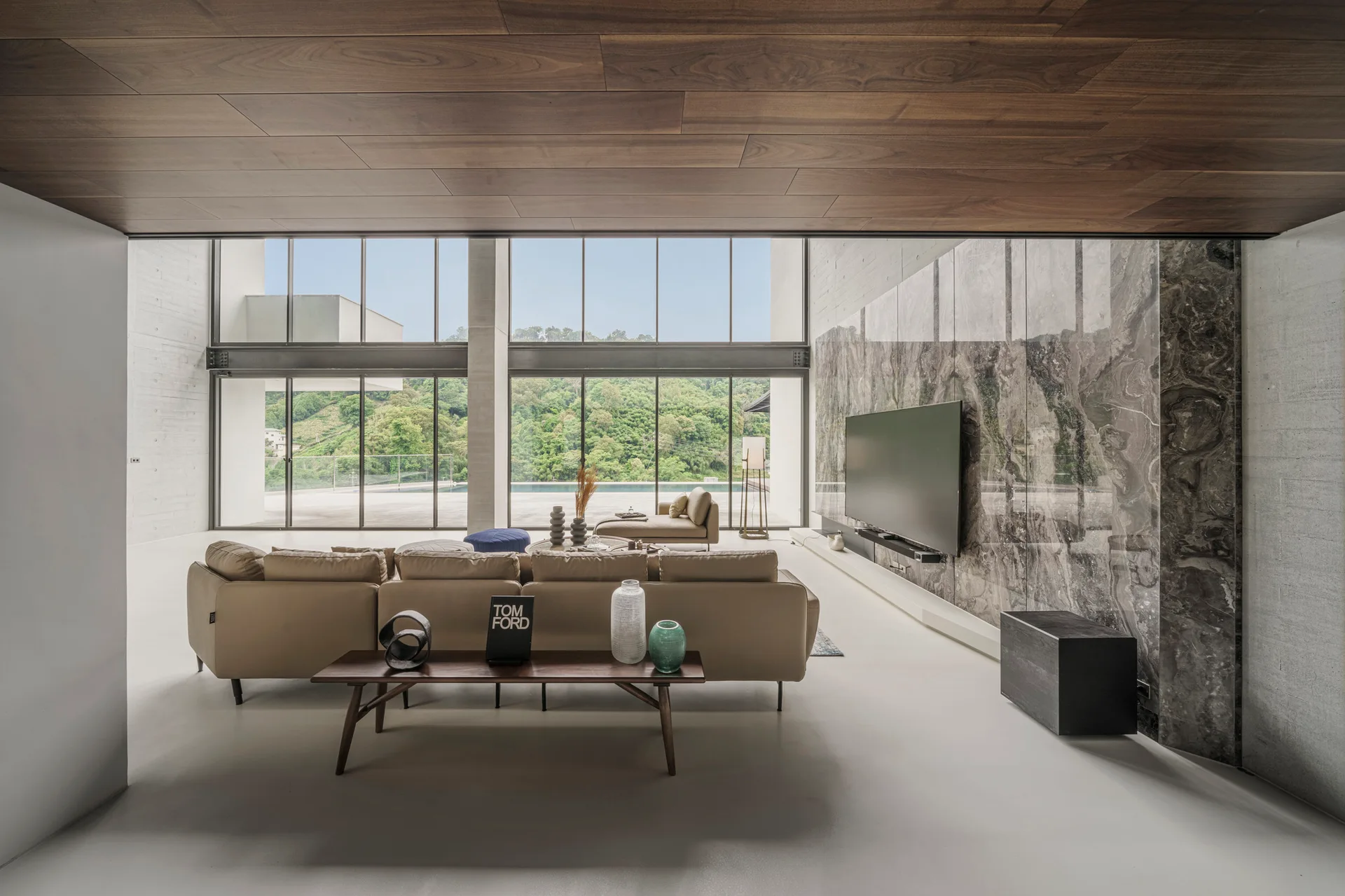 A spacious minimalist living room featuring a tan leather sectional sofa, a dark wood plank ceiling, and a large grey marble feature wall.