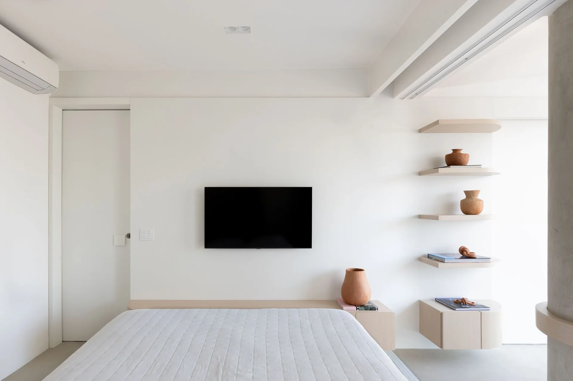 A minimalist white bedroom featuring a wall-mounted television, a white quilted bed, and light wood floating shelves styled with terracotta vases.