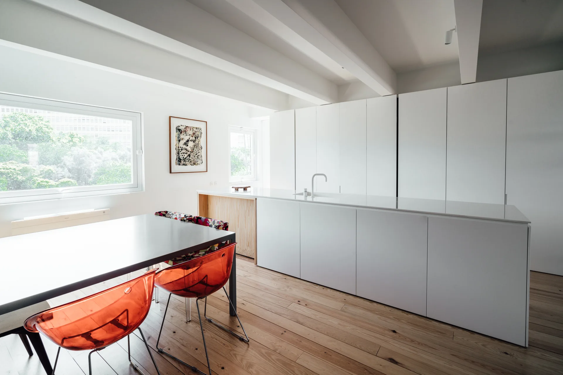 A minimalist white kitchen features seamless cabinetry, a monolithic island, light wood plank floors, and translucent orange chairs around a dining table.