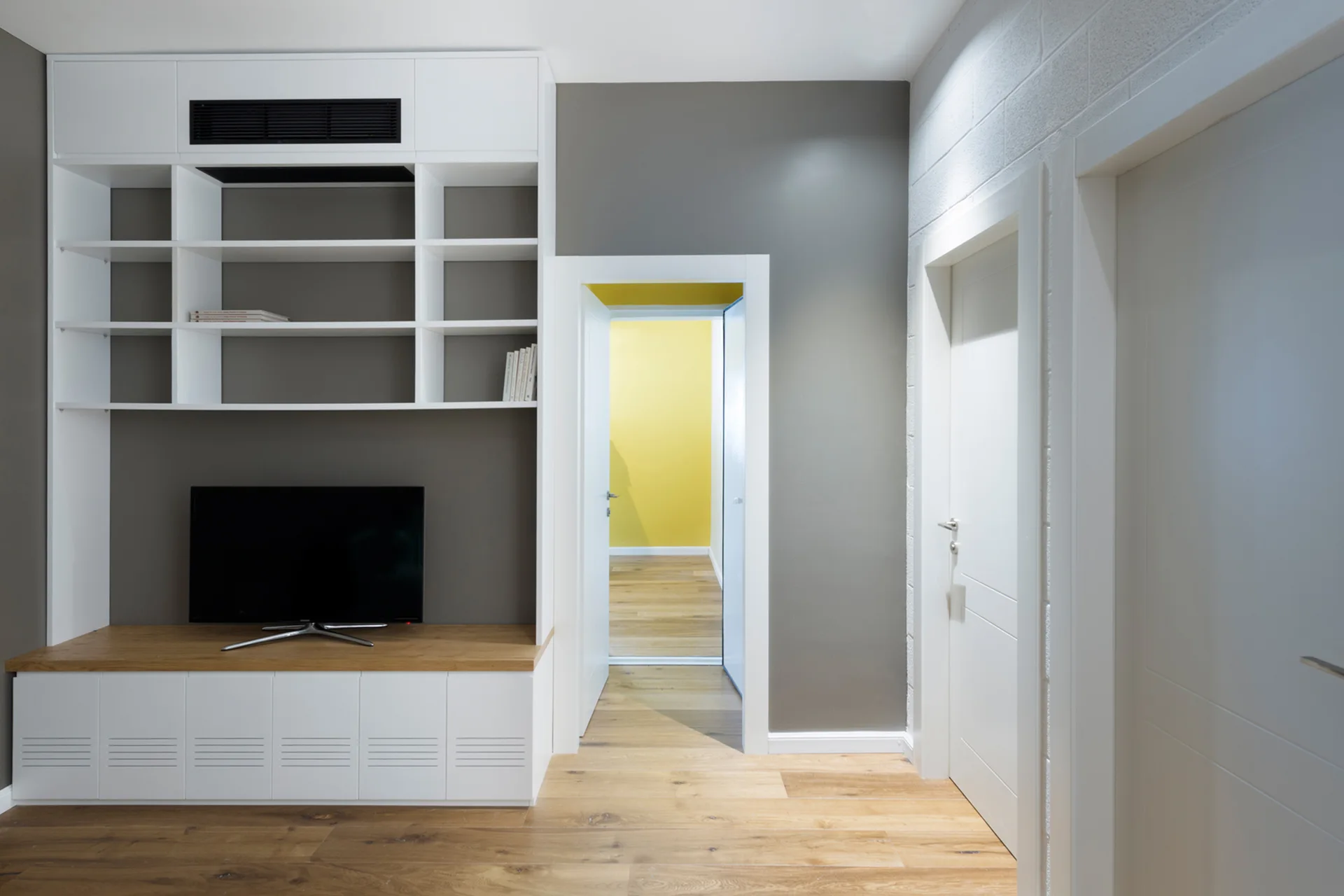 A modern hallway features a white built-in media cabinet and open shelving against a grey wall next to an open doorway showing a yellow room.