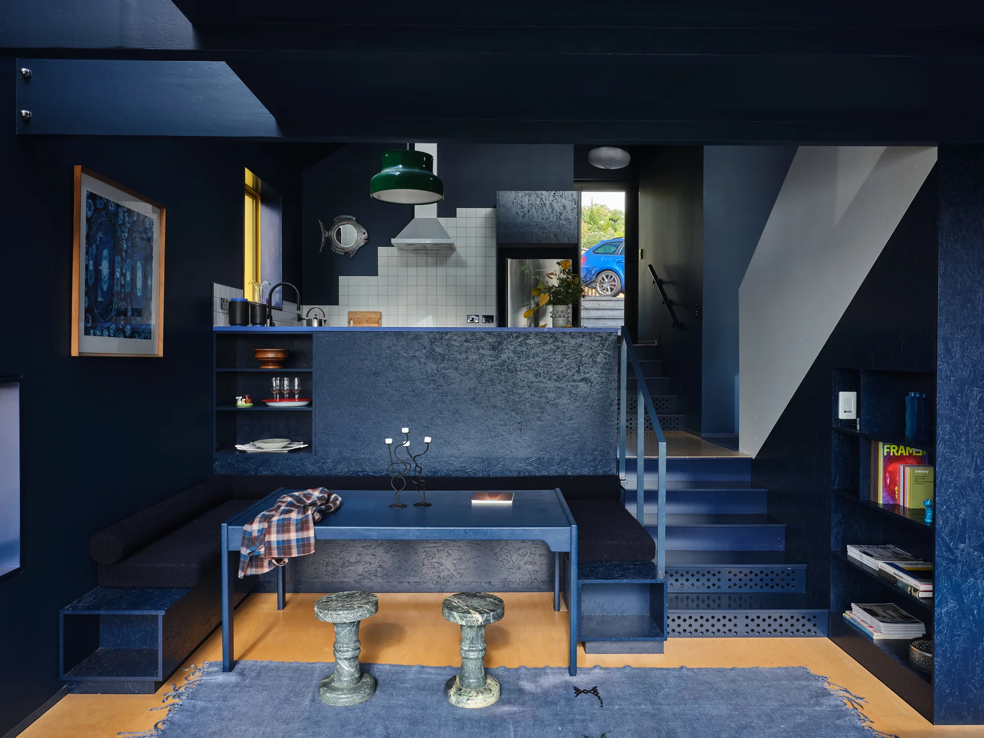 A dark blue dining nook features custom built-in bench seating, a matching blue table, green marble stools, and a staircase.