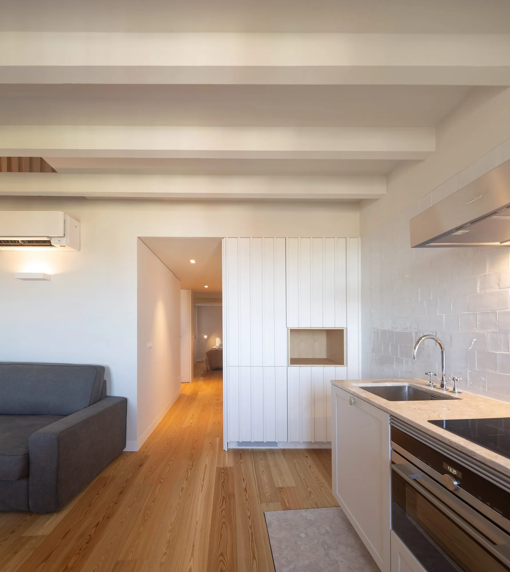 A modern open-concept kitchen features white vertical slatted cabinetry, a grey sofa, exposed ceiling beams, and light wood flooring leading to a hallway.