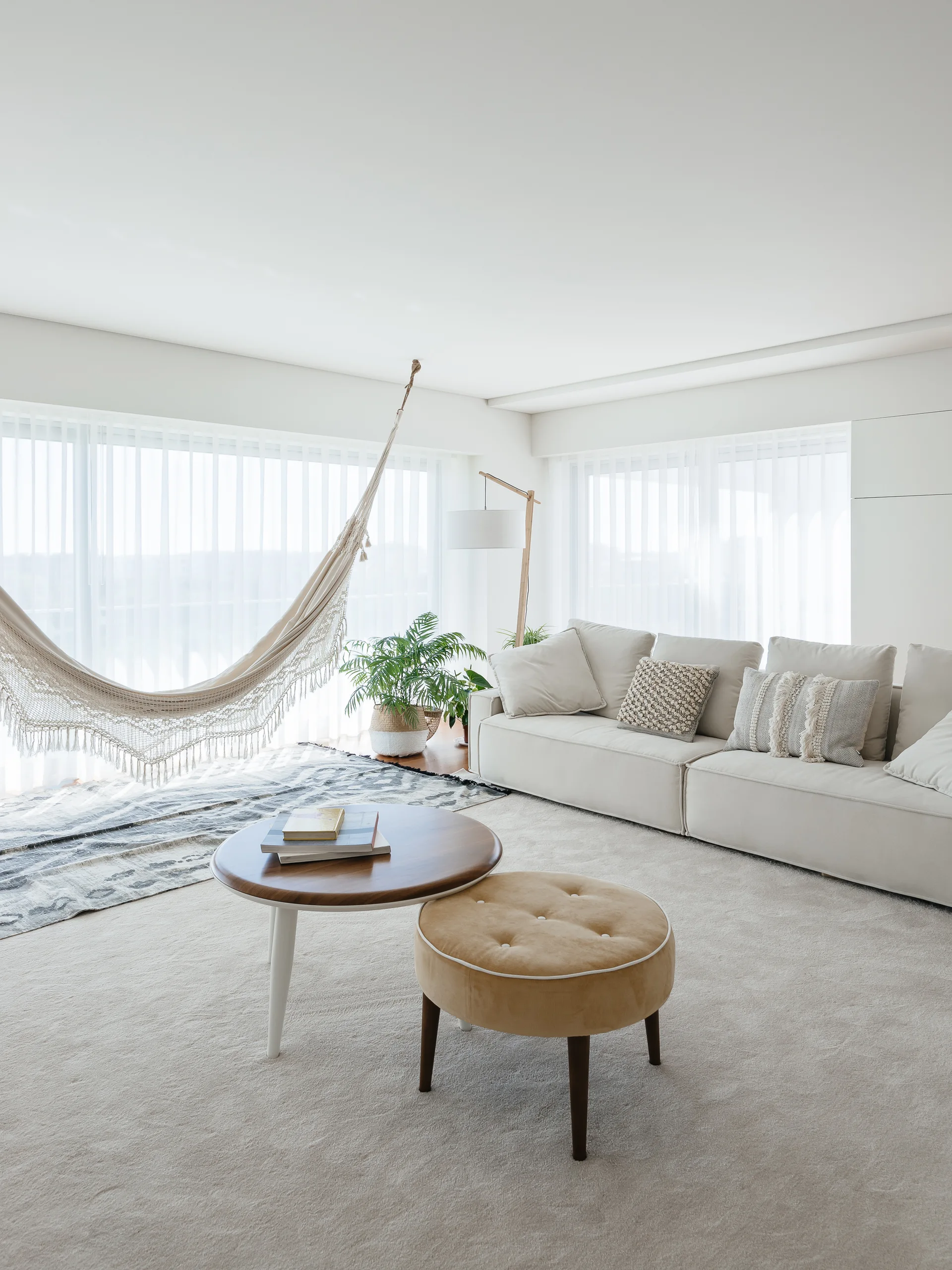 A bright neutral living room with a cream sofa, a round wooden coffee table, a tufted mustard ottoman, and a fringed woven hammock hung near the window.