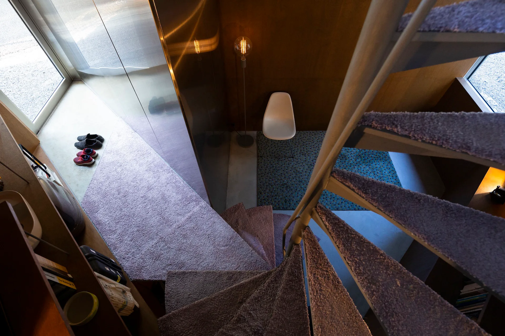 Looking down a carpeted spiral staircase into a modern entryway featuring a white shell chair, patterned blue rug, and wood-paneled walls.