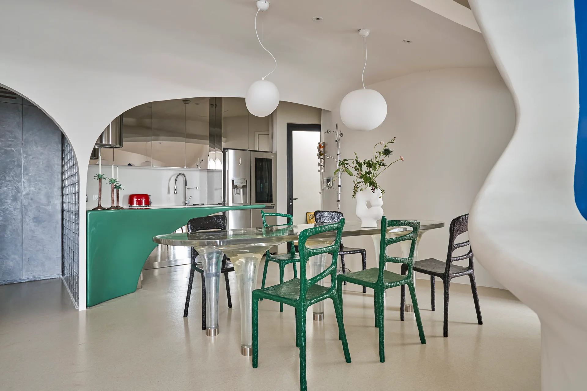 A modern dining room features a translucent sculptural table, textured green and black chairs, and a curved green kitchen counter visible through an archway.