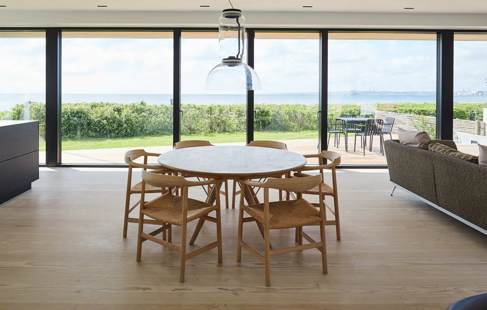 A minimalist dining area features a round light wood table and six chairs with woven seats set against large floor-to-ceiling glass walls.
