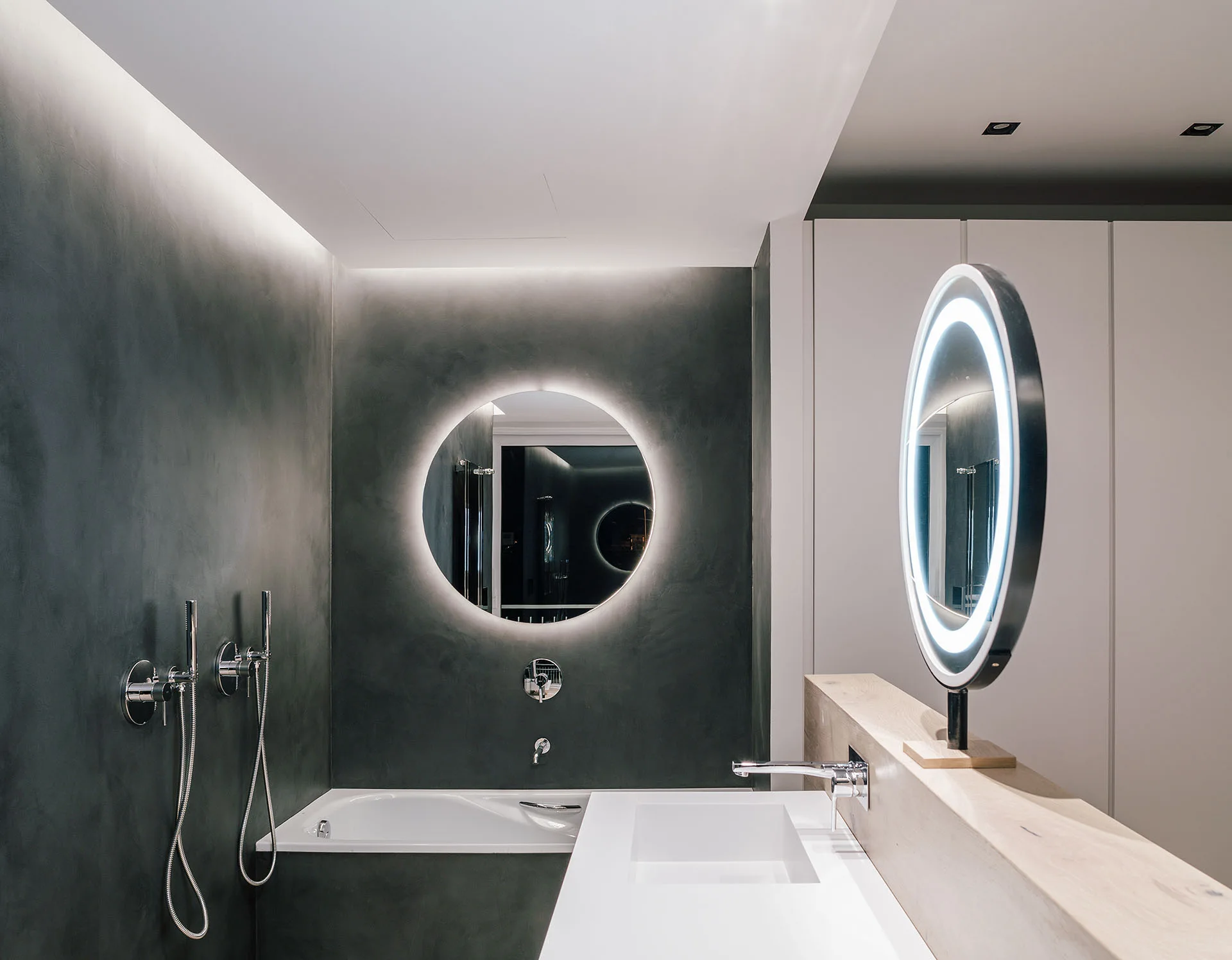 A modern bathroom features textured gray walls, a white bathtub, a wood-toned vanity counter with an integrated sink, and circular backlit mirrors.