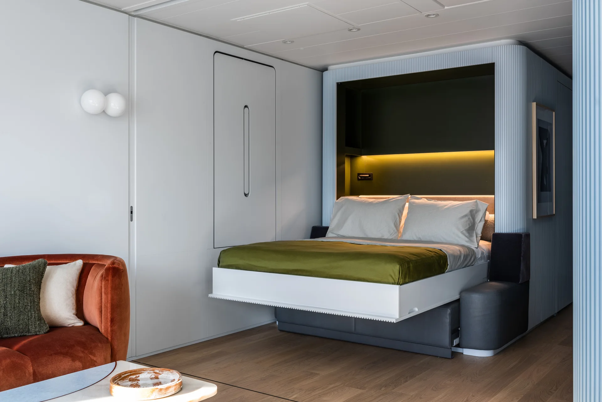 A modern room features a wall-integrated Murphy bed lowered over a grey sofa base, flanked by fluted wall paneling and a curved rust-colored sofa.