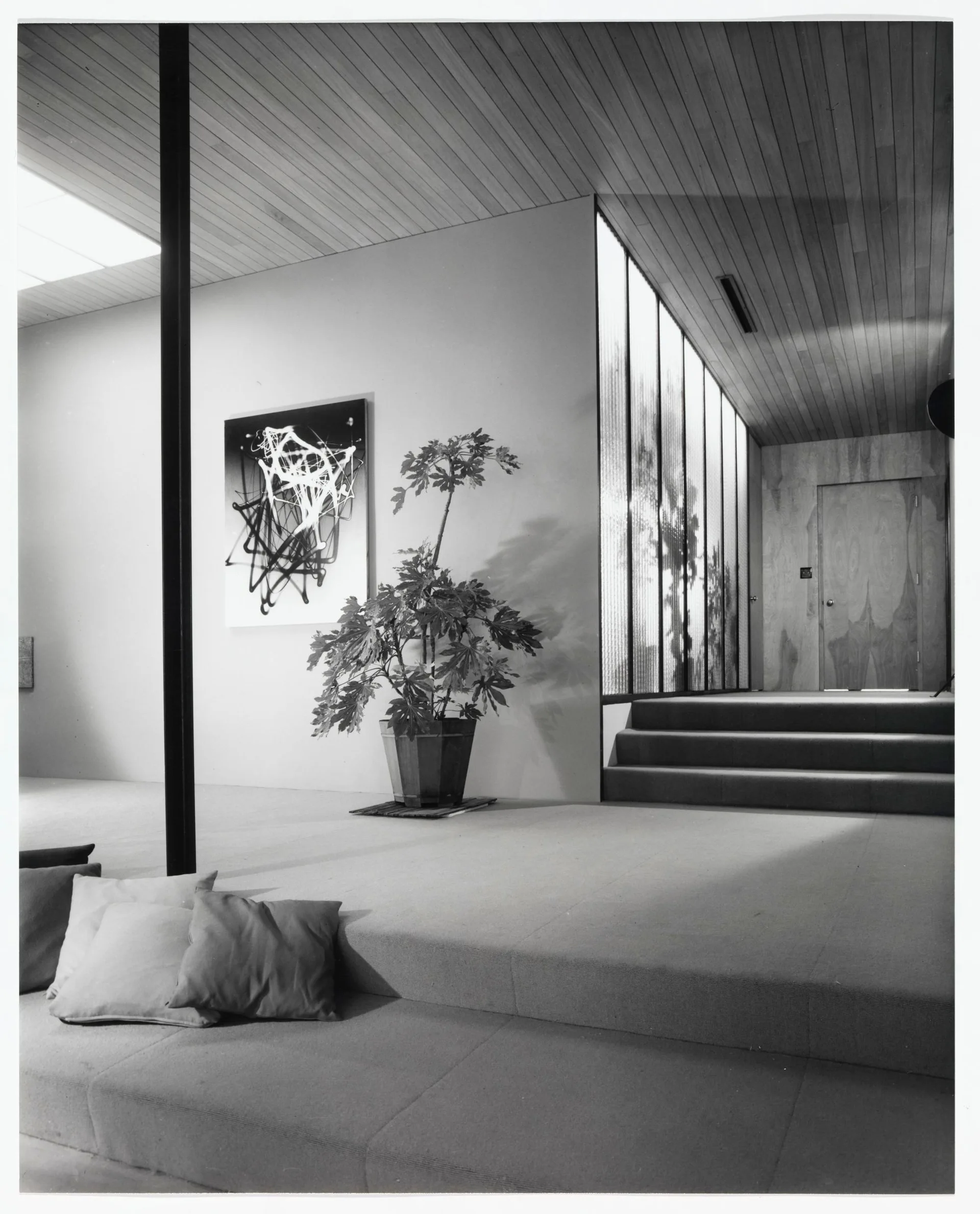 A black and white photograph of a mid-century modern sunken living room featuring a slatted wood ceiling, a fluted glass partition, and carpeted steps.