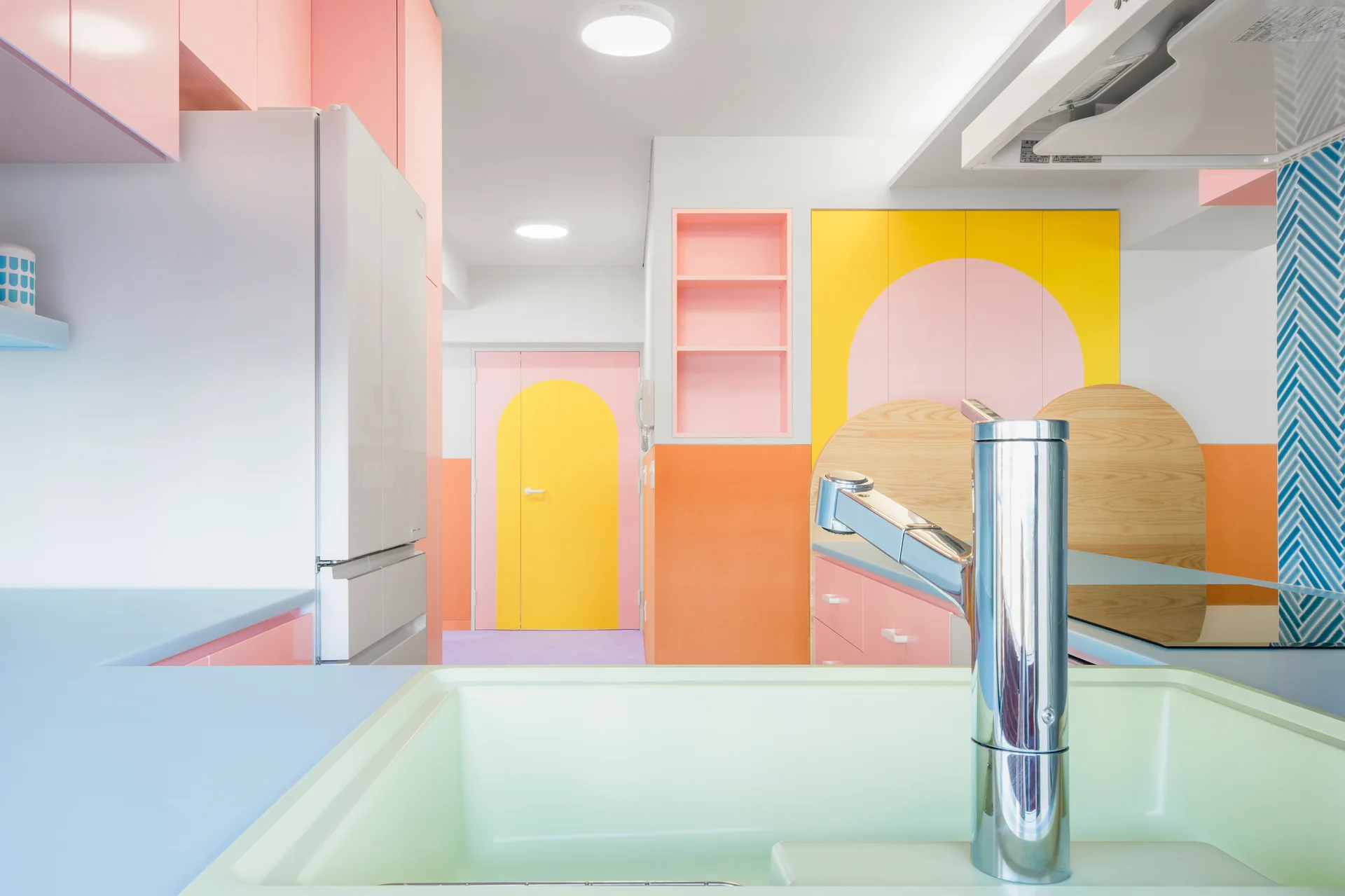 A colorful kitchen features a green sink and chrome faucet in the foreground, pastel pink and yellow cabinetry, and an arched door in the background.