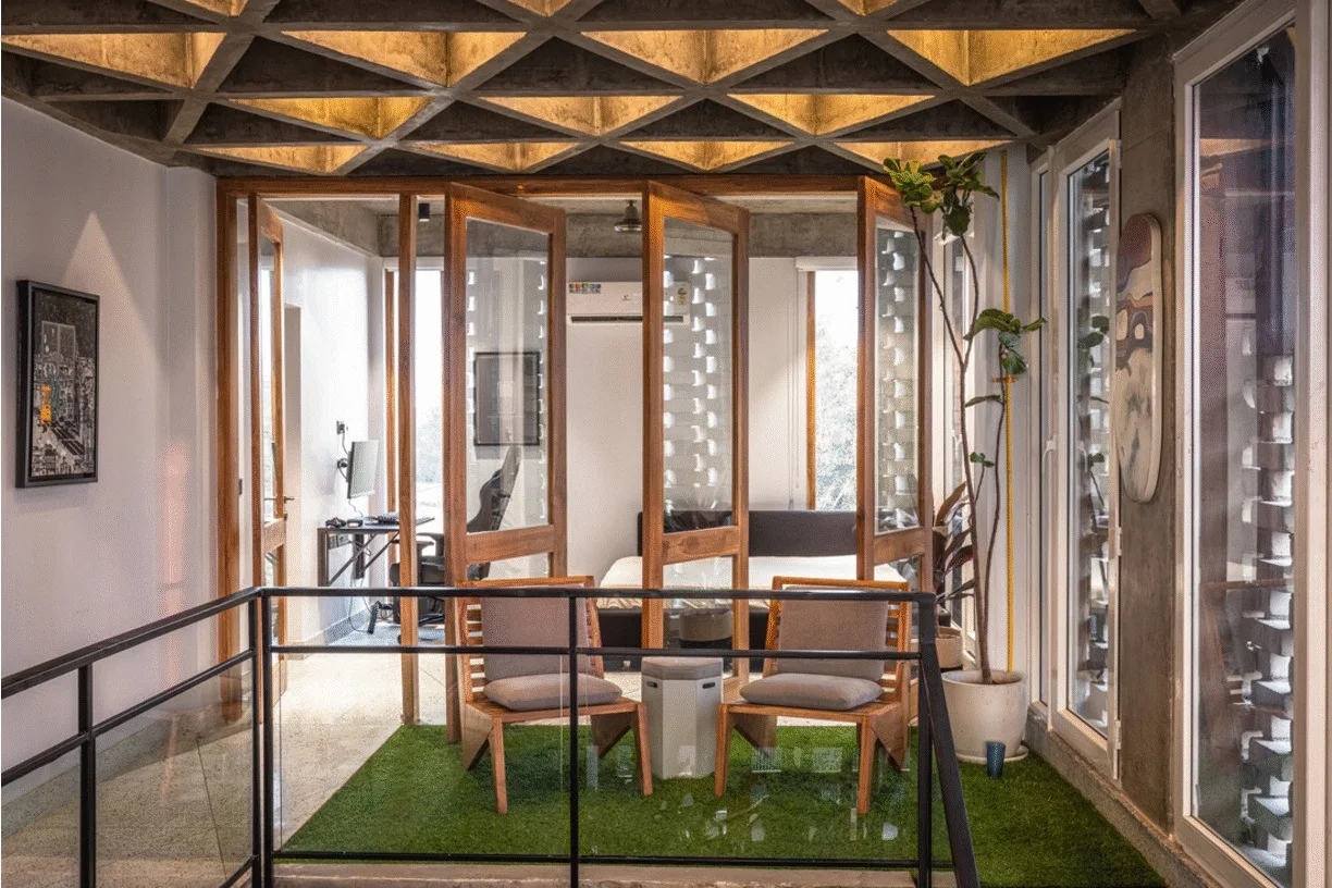 An indoor mezzanine seating area features two wooden lounge chairs on artificial turf beneath a geometric, coffered concrete ceiling.