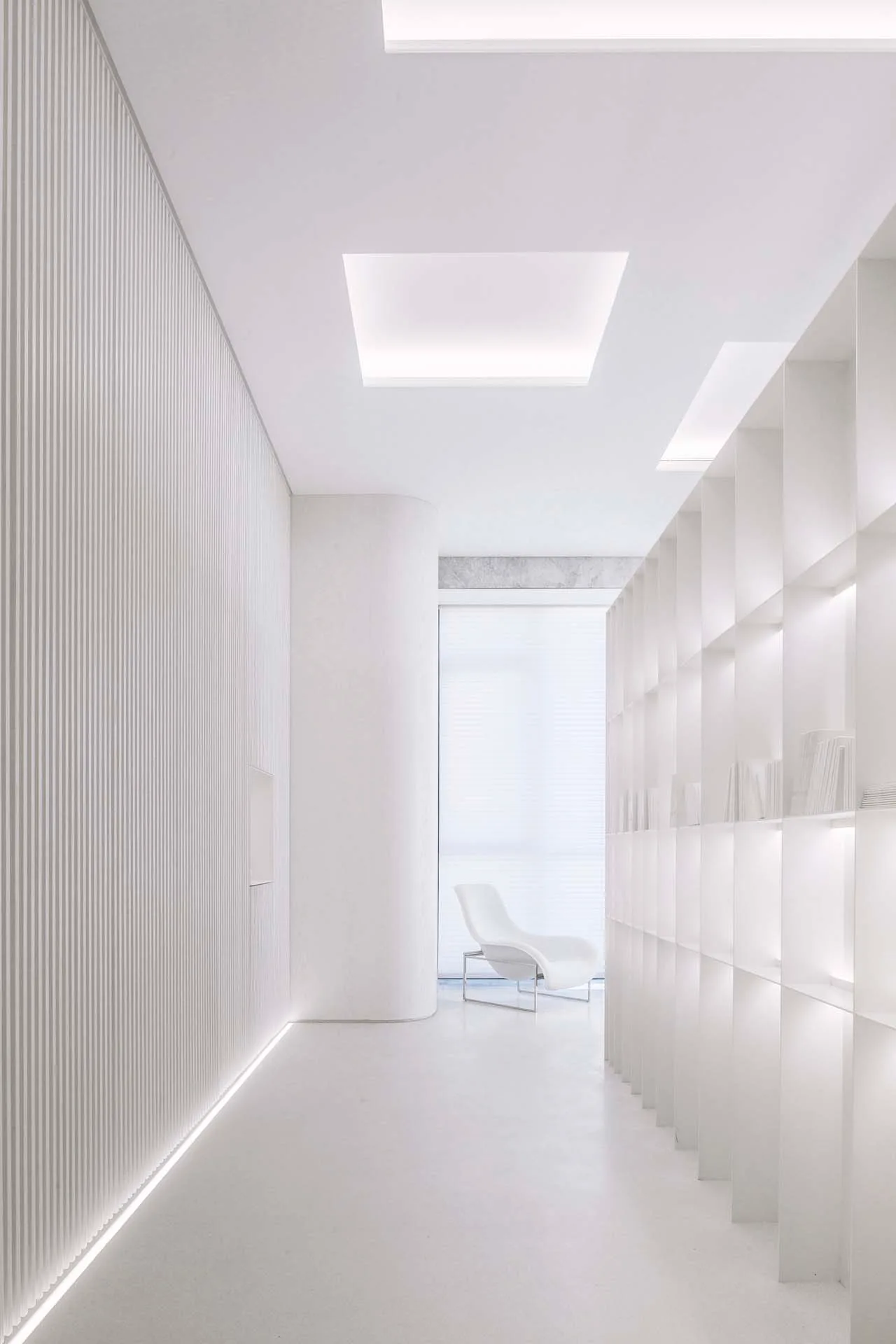 A minimalist white hallway features a vertical fluted wall, integrated grid shelving, and a modern armchair by a bright window.