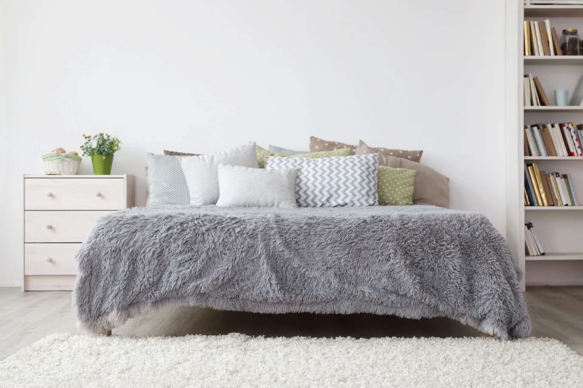 A bedroom features a bed with a fluffy grey blanket and patterned pillows next to a light wood dresser and a white bookshelf.