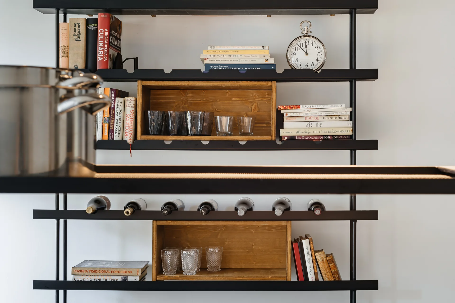 A black metal open shelving unit mounted on a white wall displays books, glasses inside wooden boxes, and horizontally stored wine bottles.