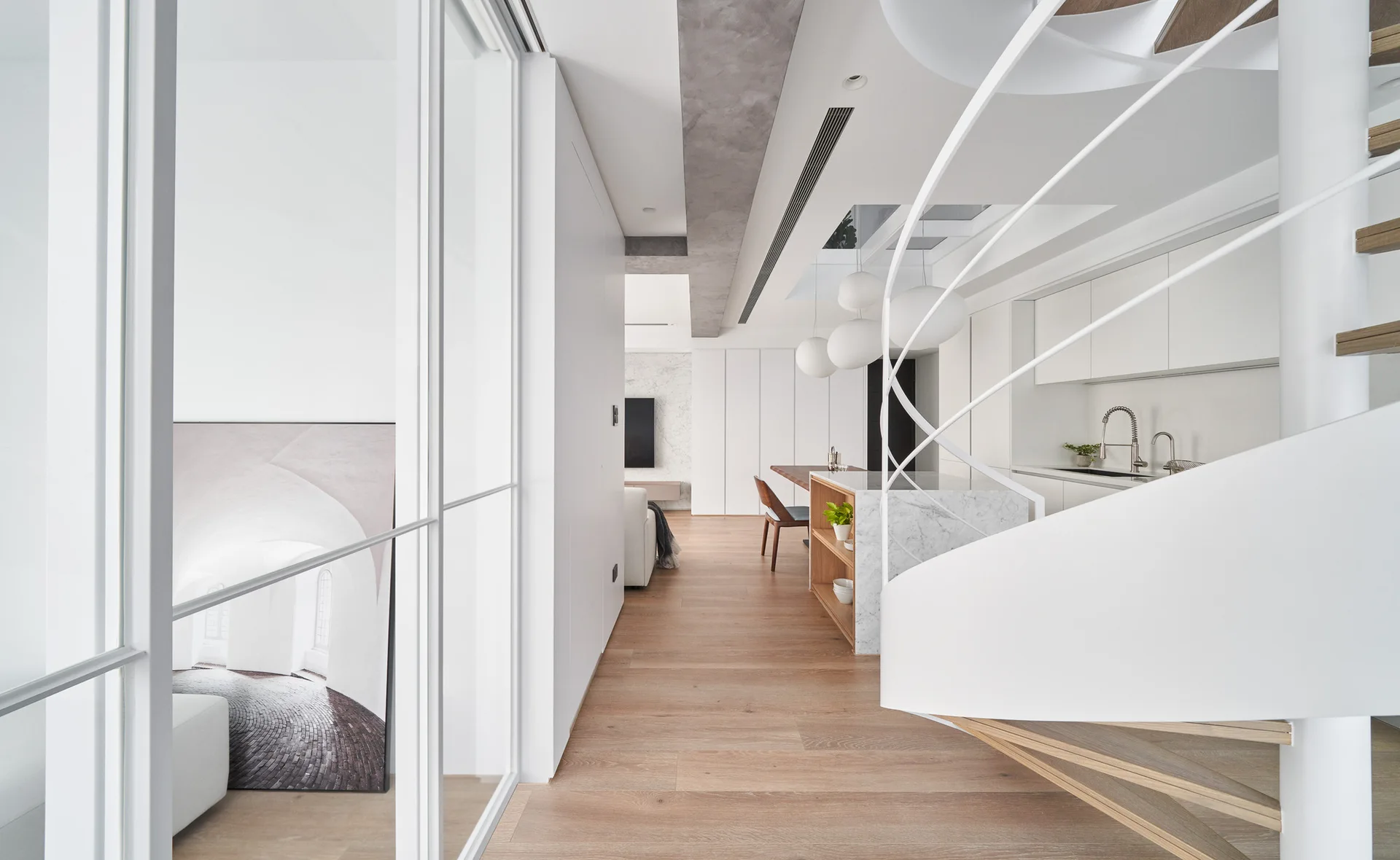A bright modern interior features a white spiral staircase in the foreground, light wood flooring, a marble kitchen island, and white handleless cabinets.