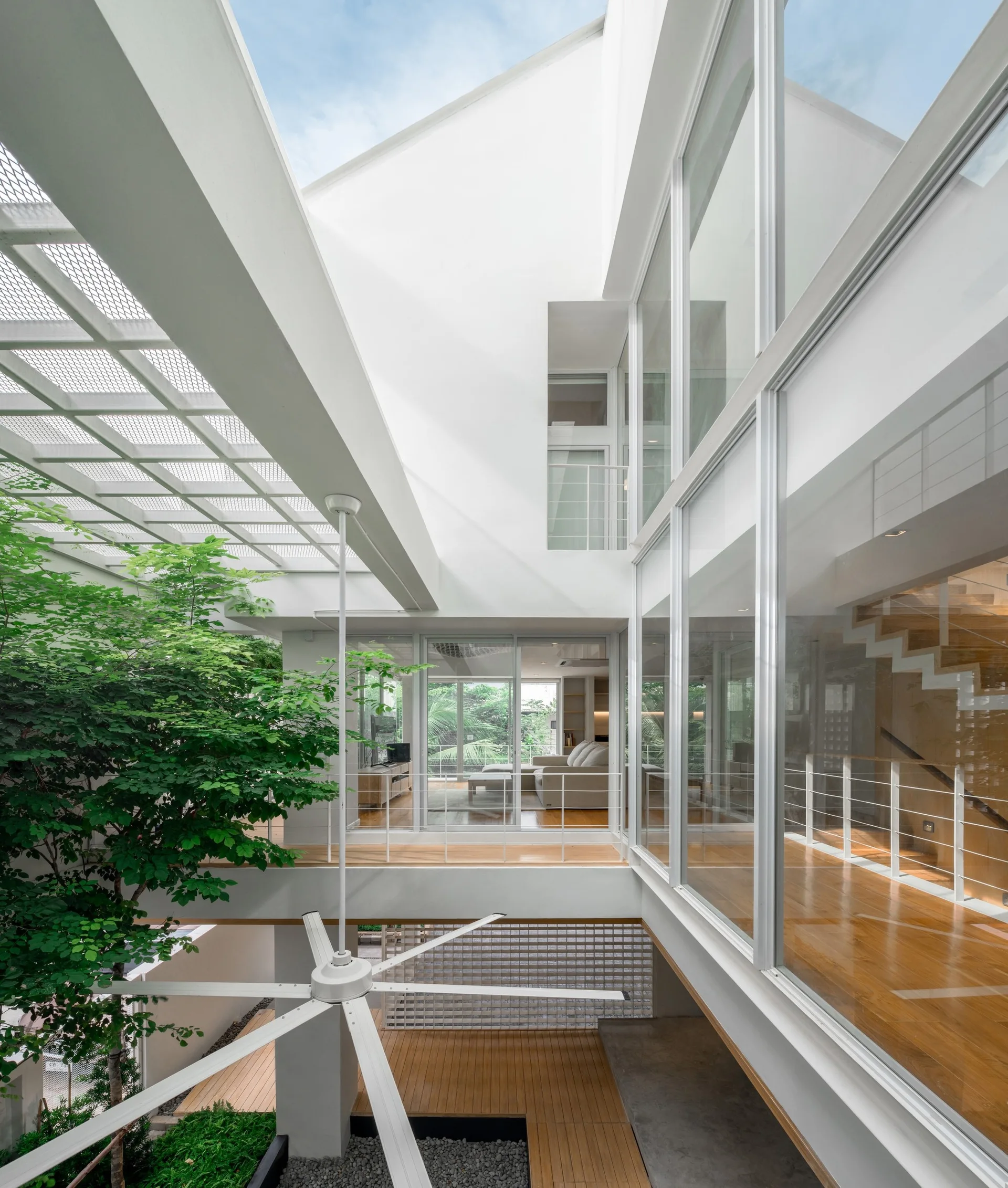 A modern multi-level house features a central glass courtyard with an indoor tree, wood flooring, and a large white ceiling fan.