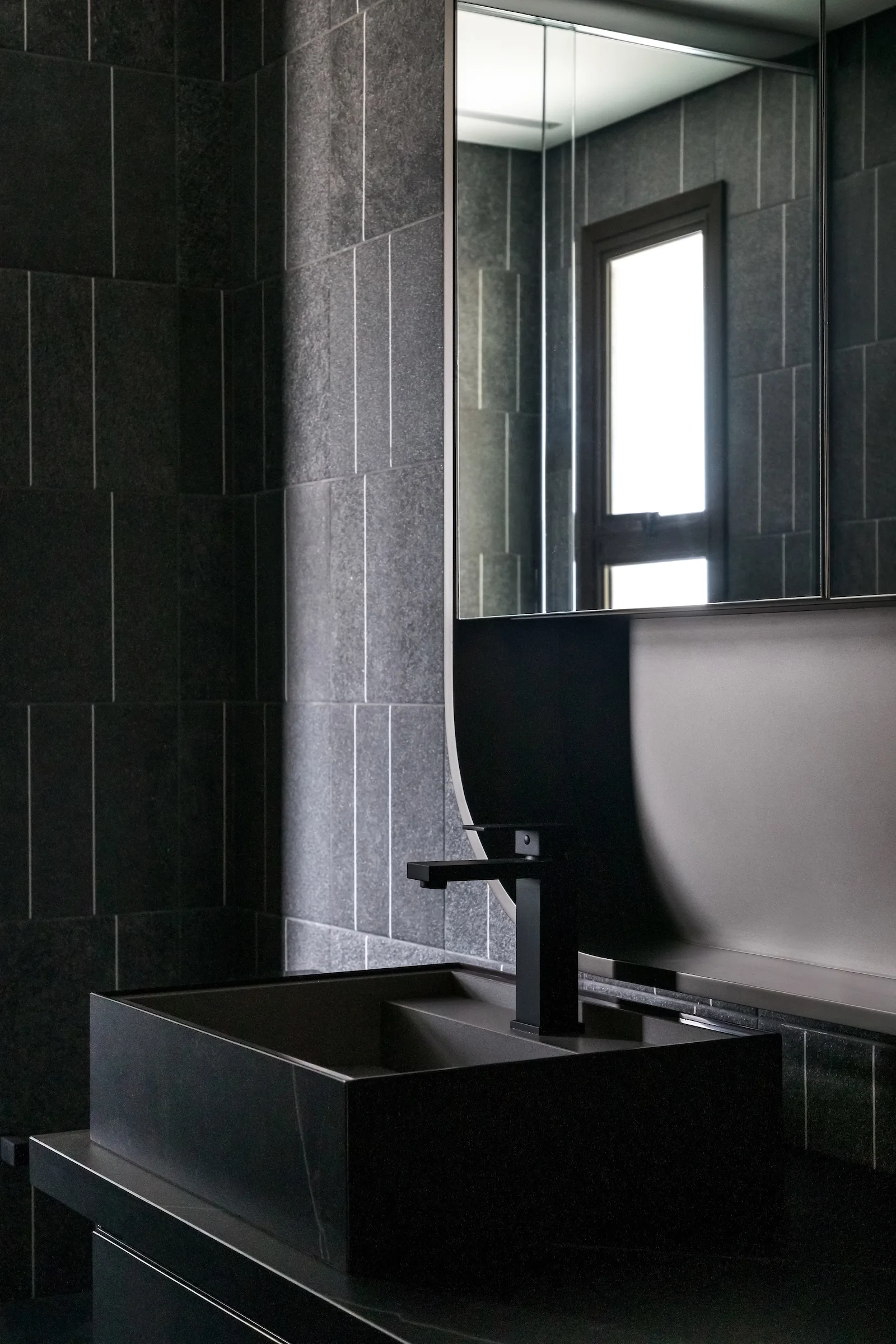 A contemporary bathroom vanity with a rectangular dark stone sink, a matte black faucet, and a wall of vertically stacked charcoal grey tiles.