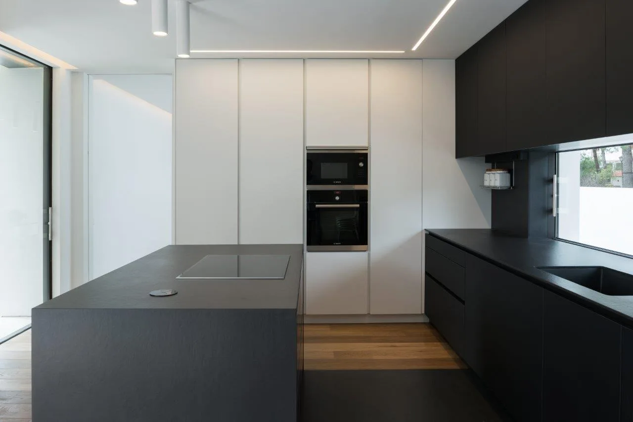 A minimalist kitchen features a matte black island, a matching black counter with a ribbon window, and tall white handleless cabinets.