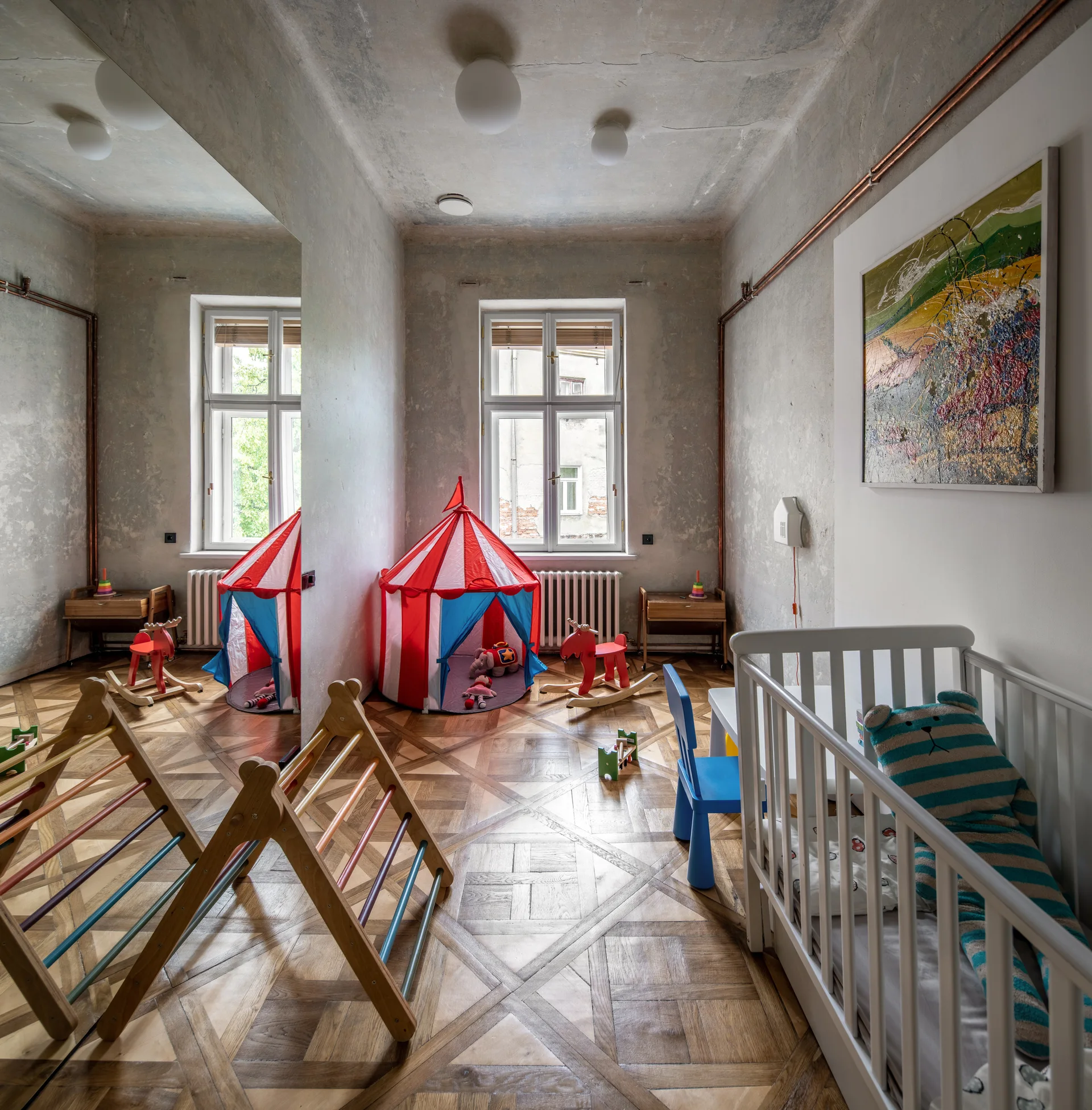 A child's room features a white wooden crib, a red and white play tent, a large wall mirror, and patterned parquet flooring under textured grey walls.