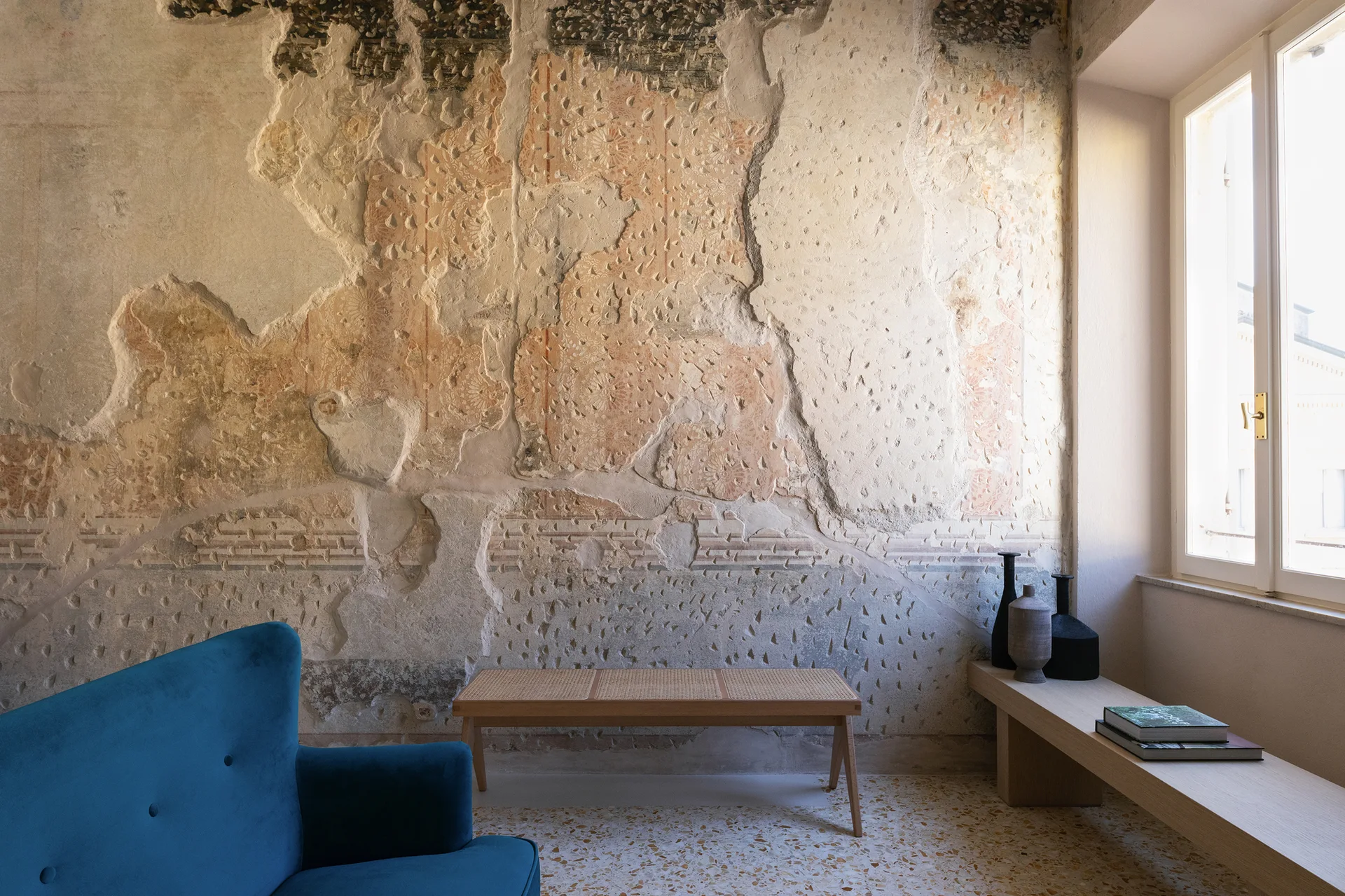 A blue velvet sofa and a light wood cane bench are set against a heavily distressed historic plaster wall in a room with a terrazzo floor.