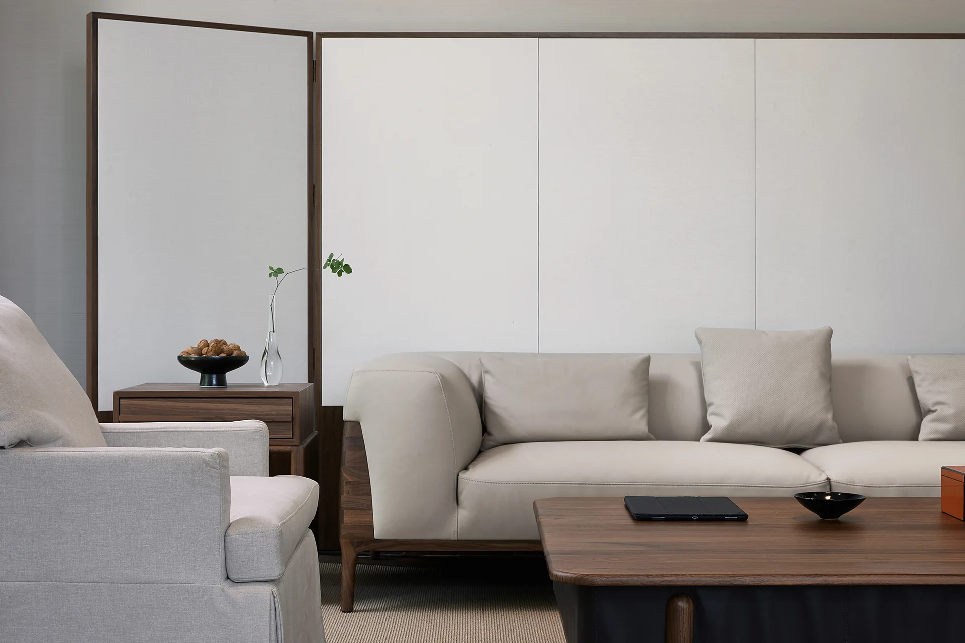 A minimalist living room with a cream-colored sofa, dark wood coffee table, grey armchair, and a large white folding screen backdrop.
