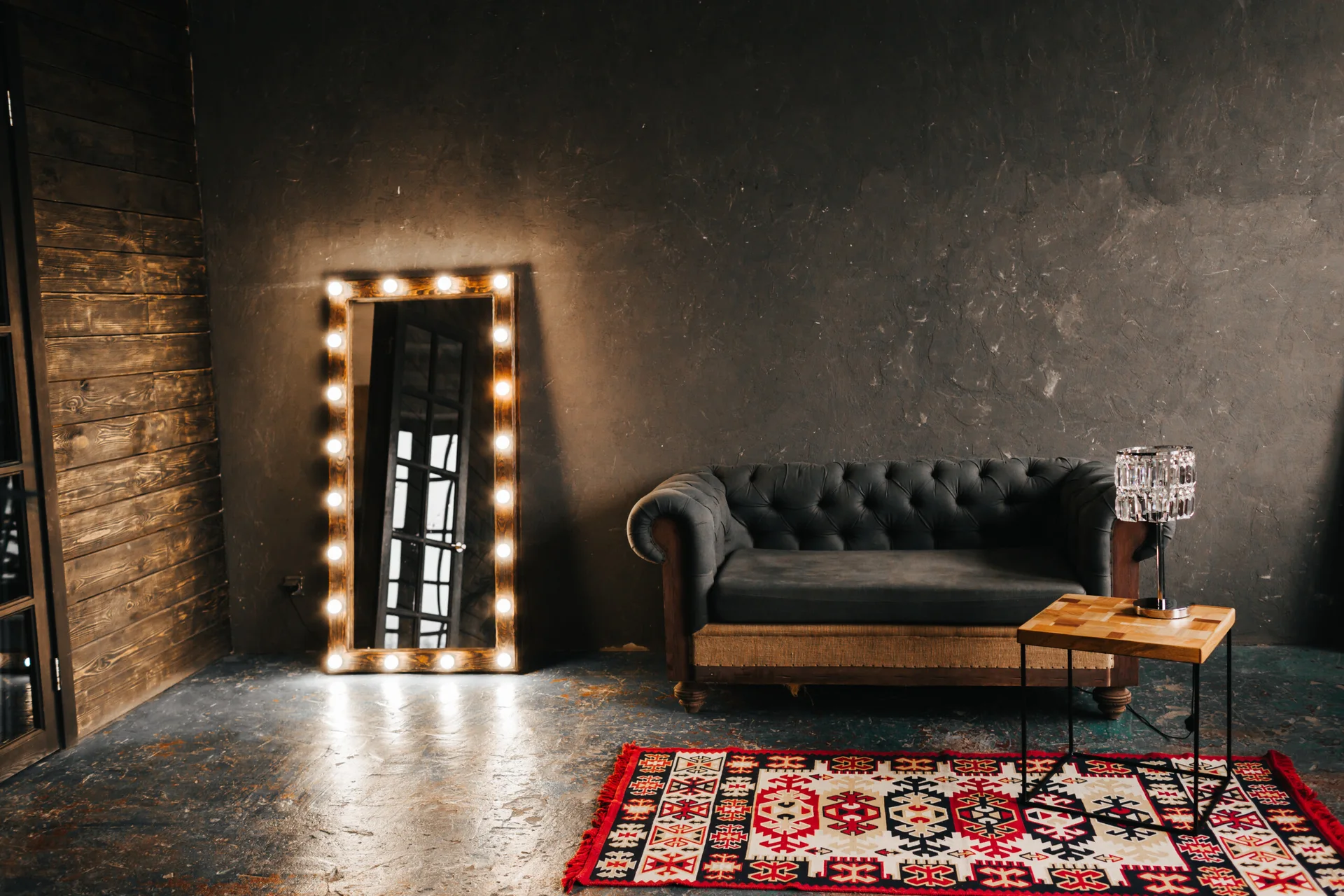 A tufted grey chesterfield sofa sits against a textured dark wall next to a large floor mirror framed in lit lightbulbs on a patterned rug.