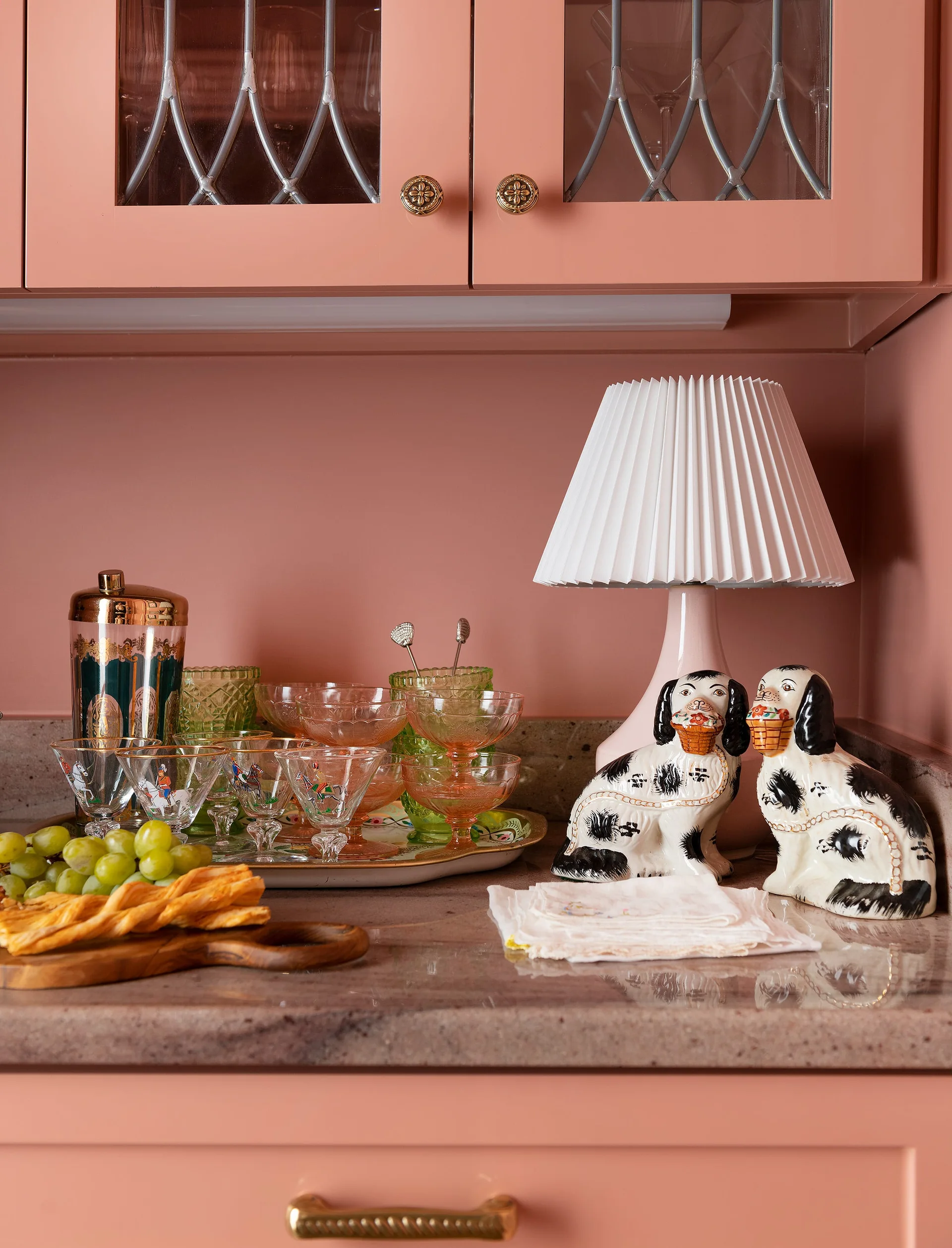 A pink kitchen countertop featuring two ceramic dog figurines, a small pleated table lamp, and a cocktail shaker tray below pink cabinets.