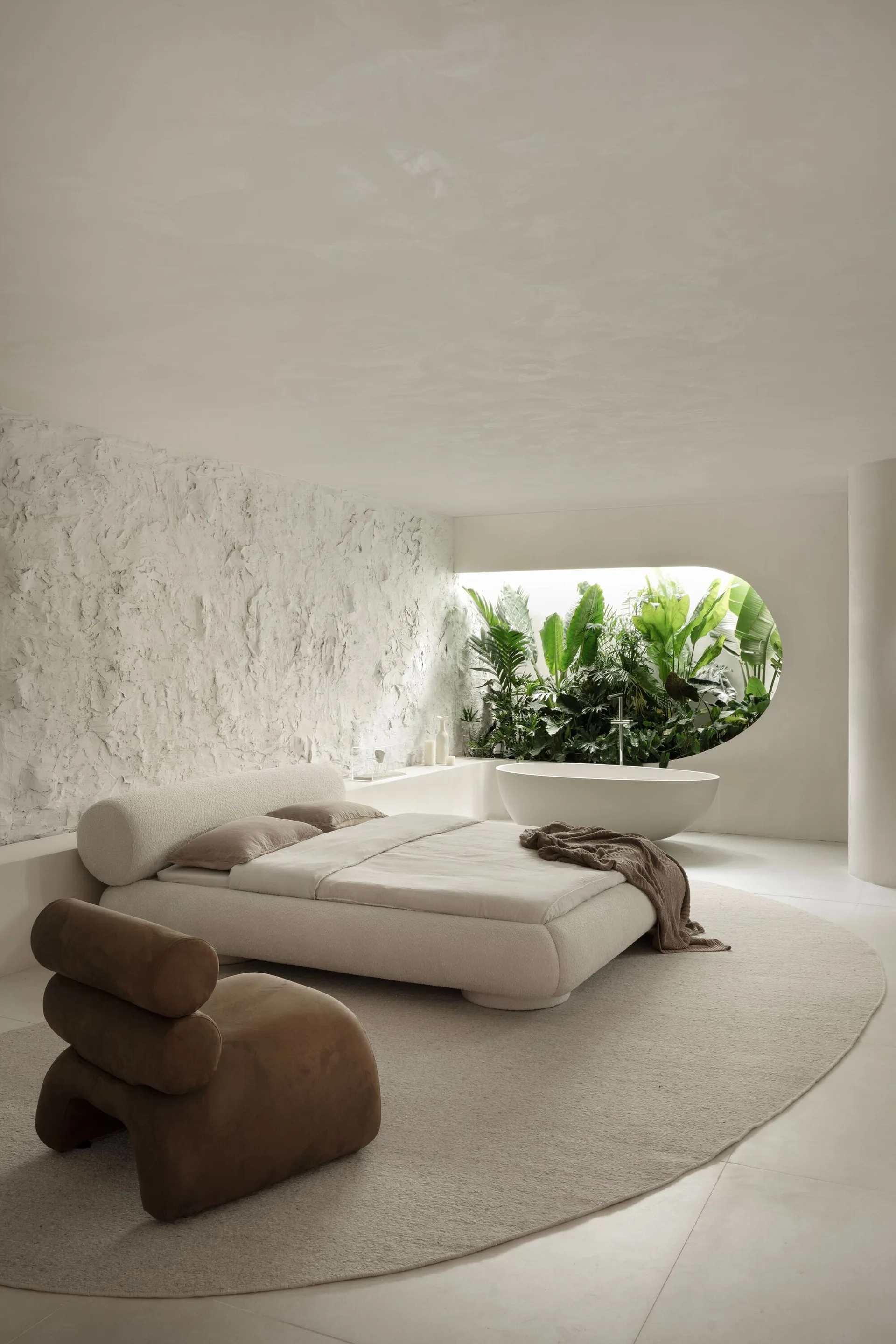 An organic modern bedroom features a cream platform bed, a brown sculptural chair, a rough plaster wall, and an oval window framing green plants.