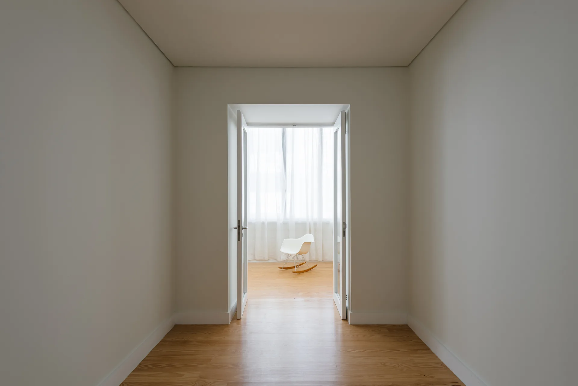 An open doorway at the end of a white hallway leads to a room with light wood floors and a white rocking chair in front of sheer curtains.