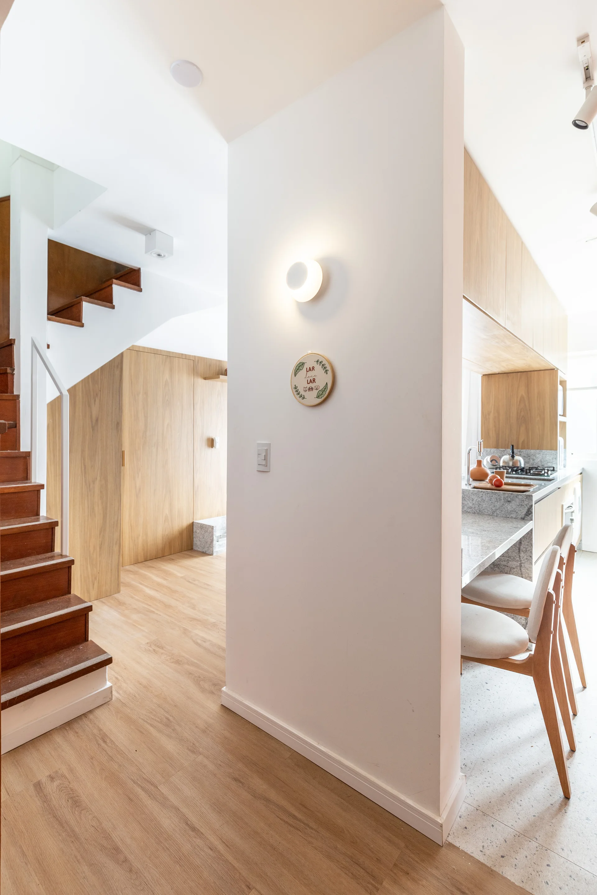 A white partition wall with a round glowing sconce separates a wooden staircase from a kitchen with light wood cabinets and two counter stools.