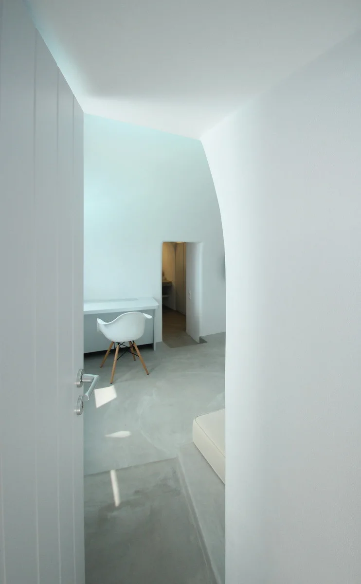 A minimalist white bedroom featuring a molded plastic chair at a desk, seamless grey flooring, and curved plaster walls viewed through a doorway.