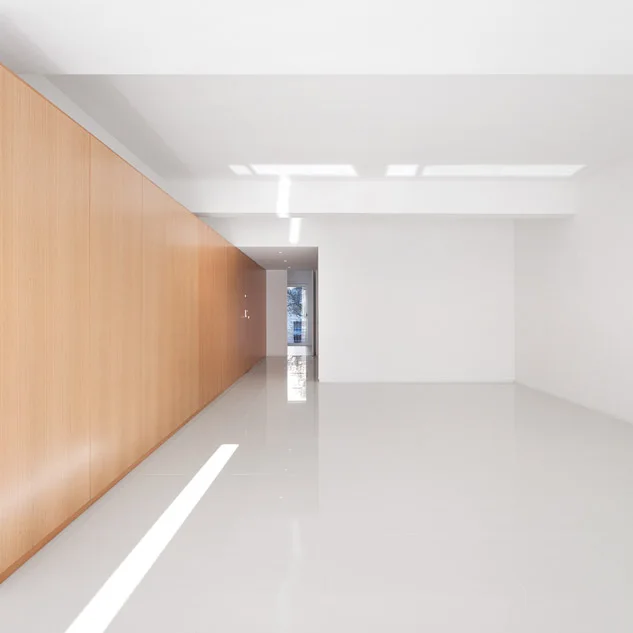 A minimalist empty hallway features a long wall clad in light wood paneling opposite a plain white wall and reflective polished white tile flooring.