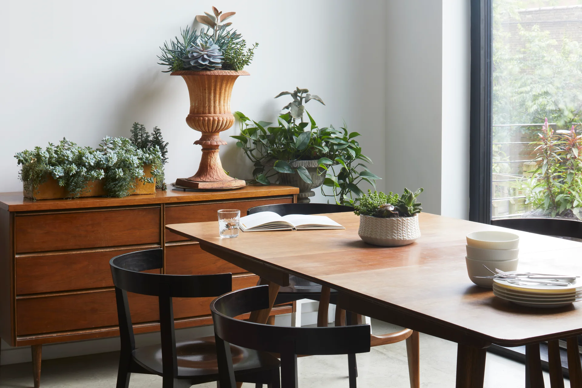 A wooden dining table with black chairs features an open book, while a mid-century credenza nearby holds planters with dense green succulents.