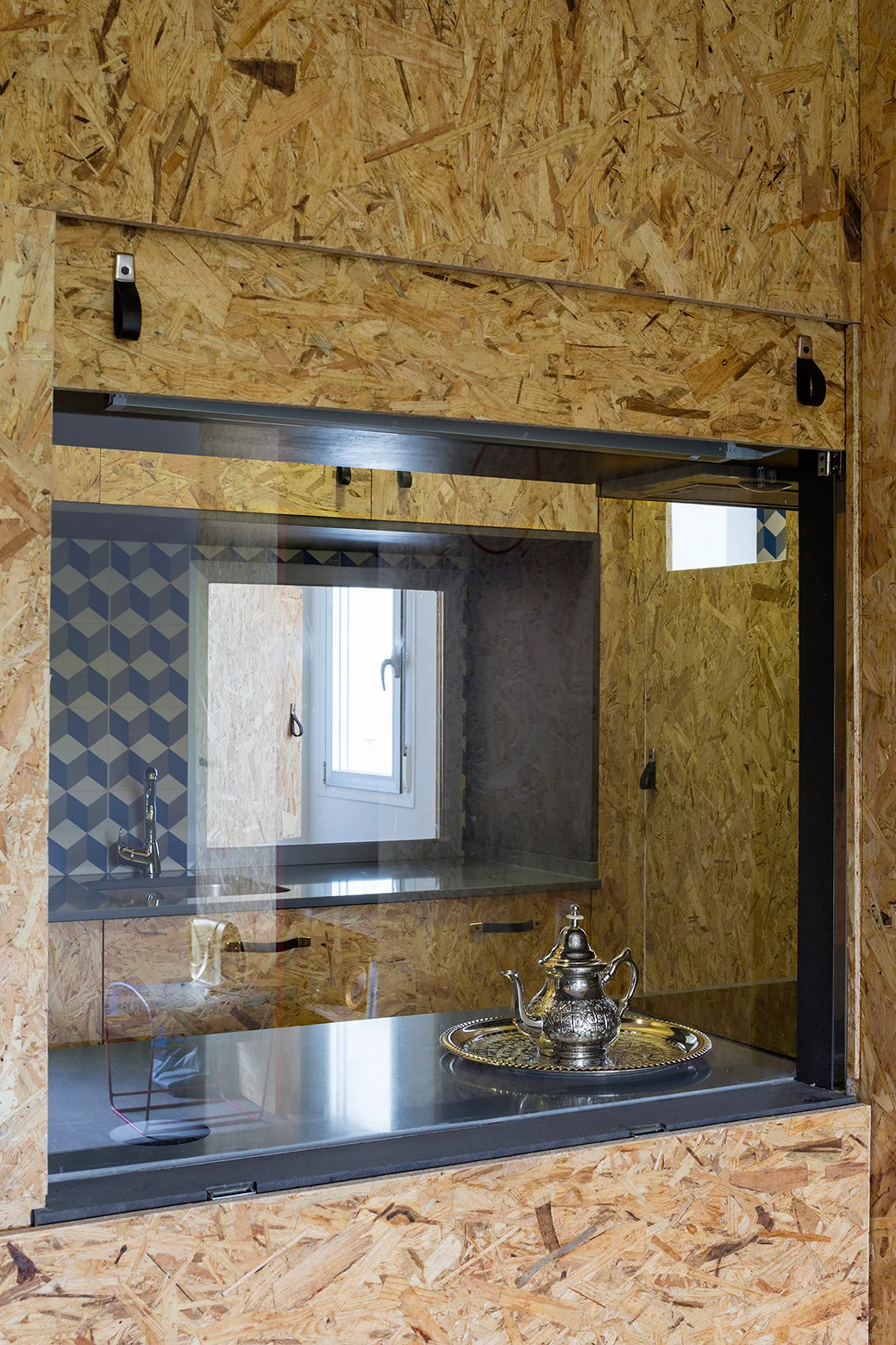 A rectangular kitchen pass-through framed in oriented strand board reveals a gray countertop, a sink, and a blue geometric patterned tile backsplash.