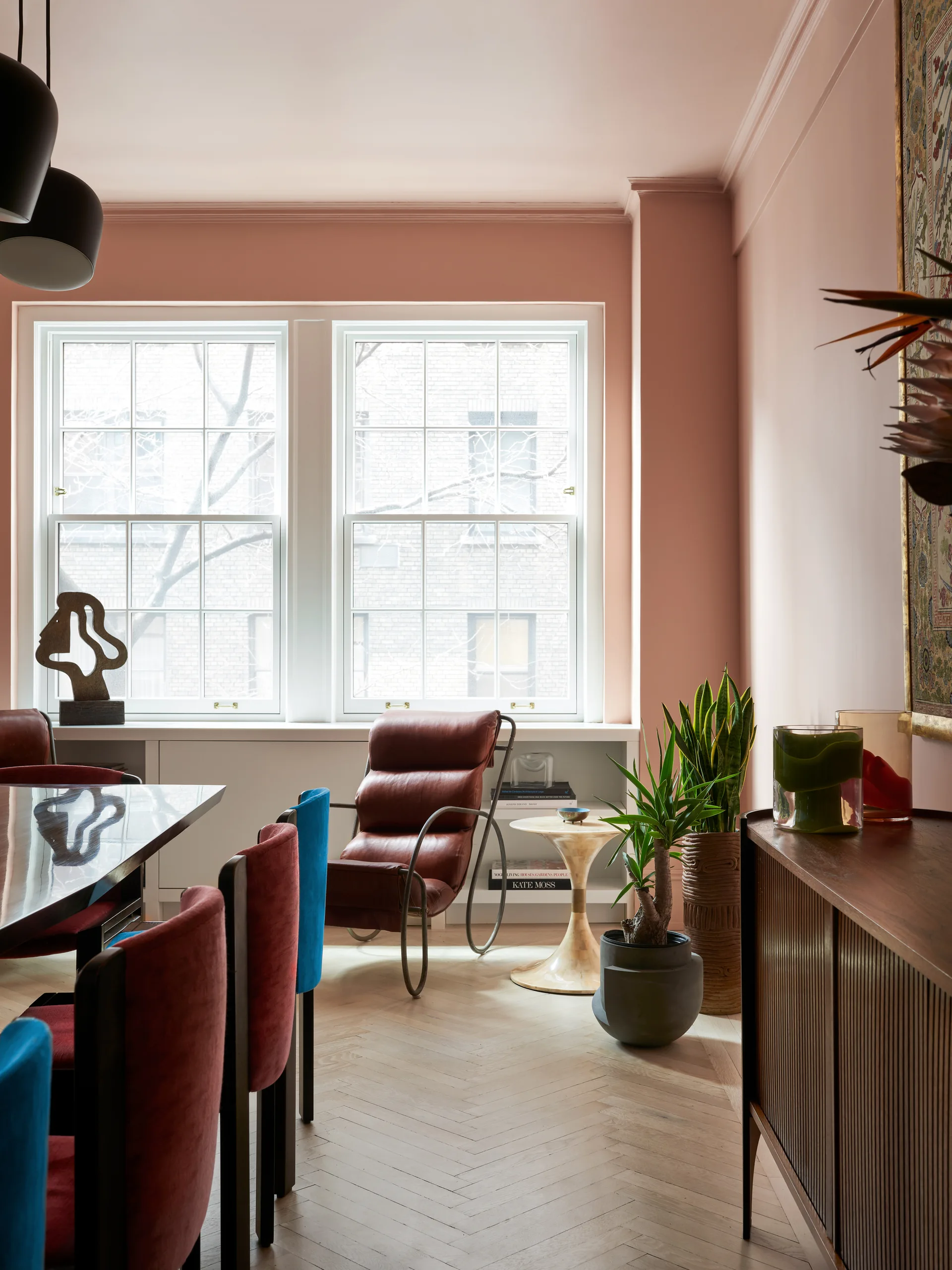 A dining room features blush pink walls, light wood herringbone flooring, a brown leather lounge chair by a window, and colorful dining chairs.