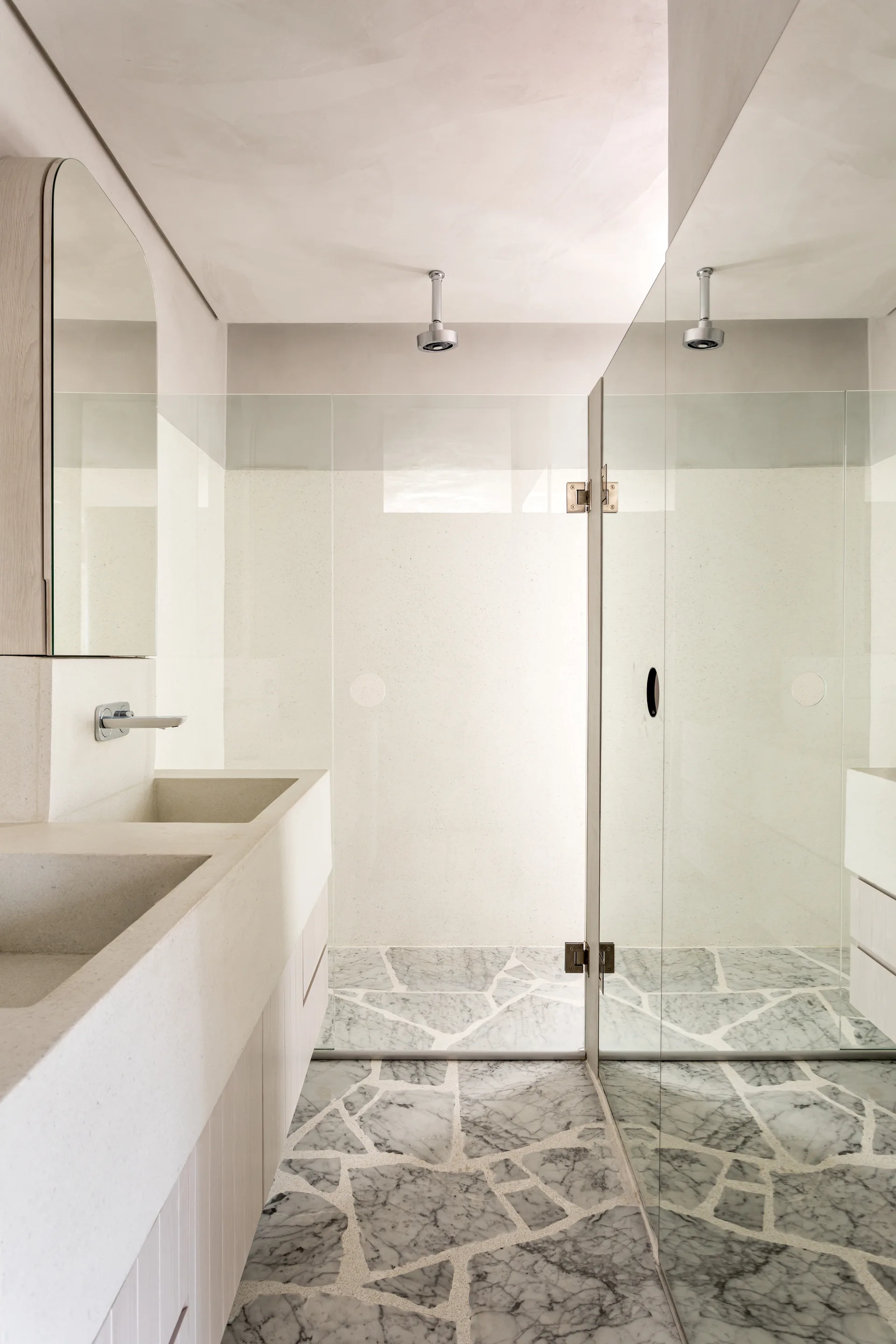A minimalist bathroom features an integrated stone double sink vanity, a glass shower, and irregular grey and white marble stone floor tiling.