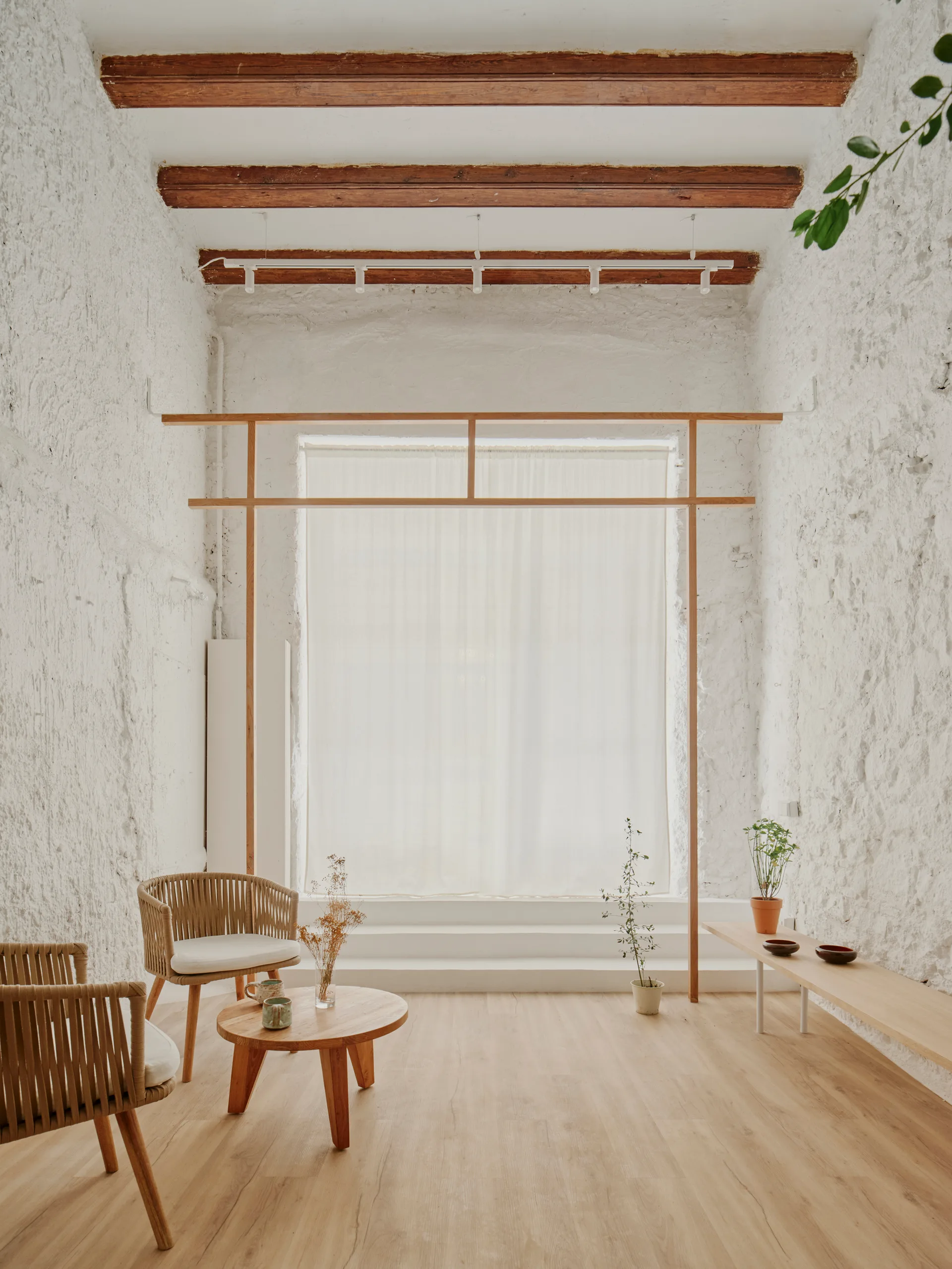 A minimalist living space featuring rough textured white plaster walls, dark wooden ceiling beams, woven armchairs, and a low wooden bench.