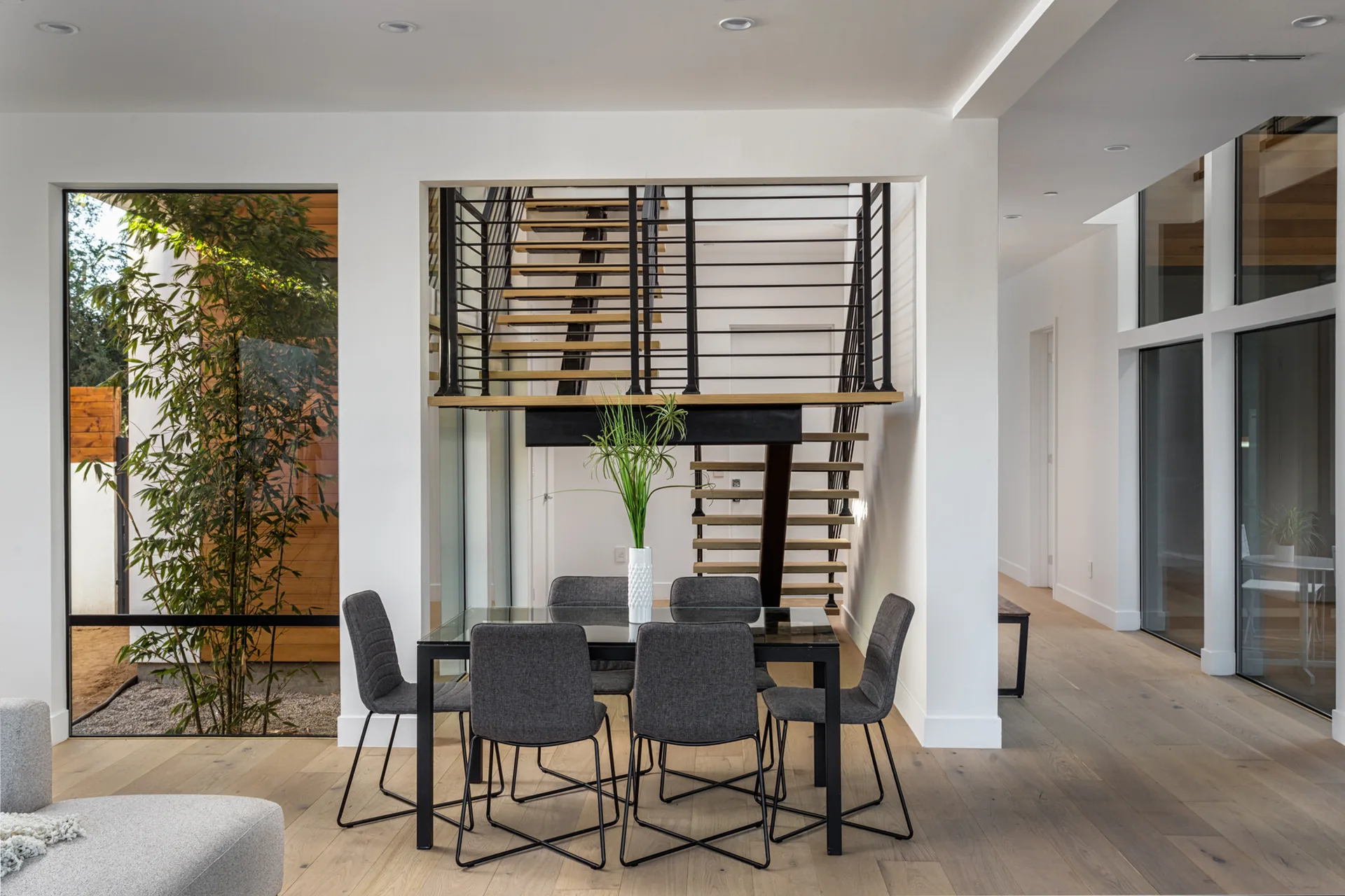 A modern dining room features a glass table with six gray chairs set before an open-riser wooden staircase and a tall window framing green bamboo.