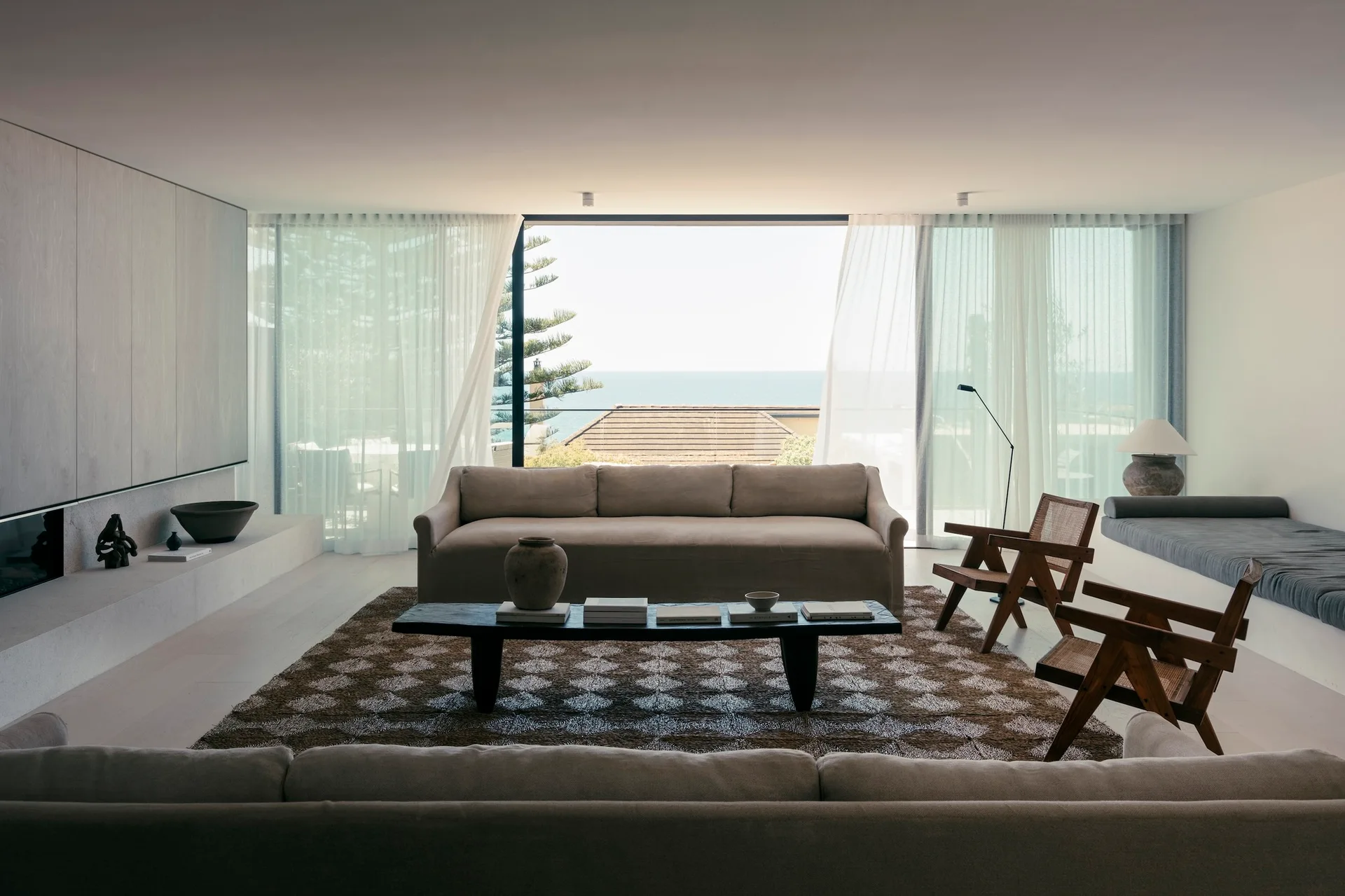 Sheer white curtains frame a large glass door overlooking the ocean in a minimalist living room with beige sofas and a low dark wooden coffee table.