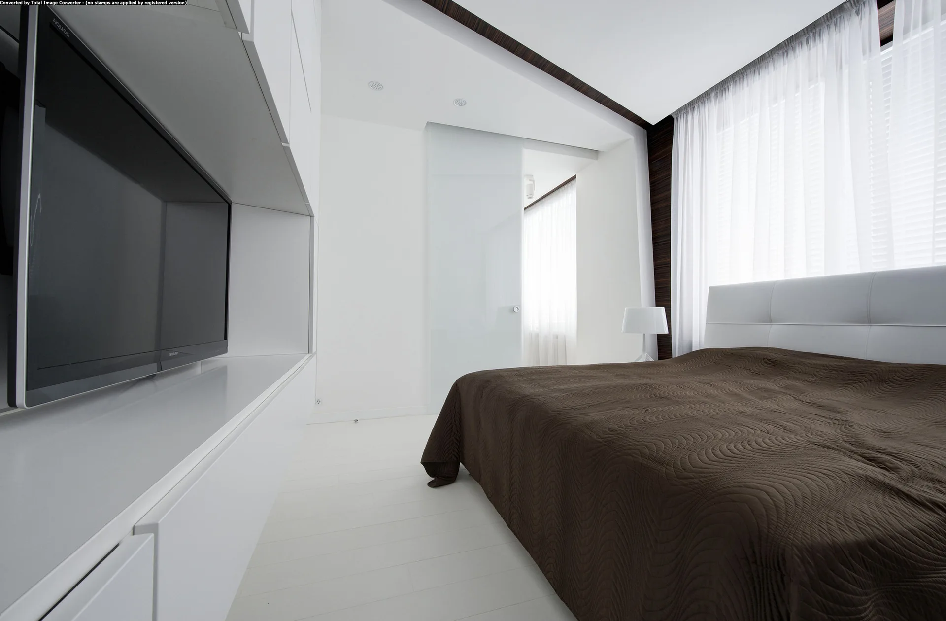 A minimalist white bedroom features a bed with a dark brown quilt, a white headboard, a wall-mounted TV in built-in cabinets, and sheer curtains.