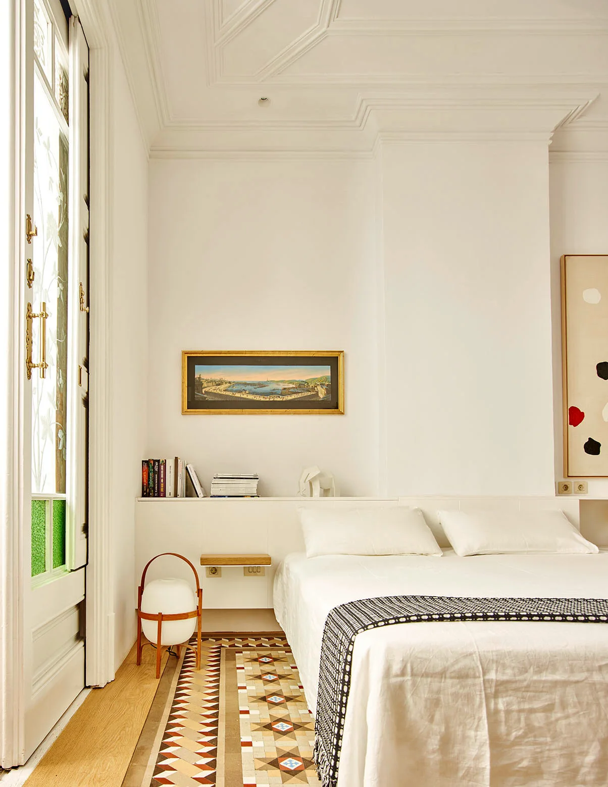 A bright white bedroom features a bed with white bedding, a built-in headboard ledge holding books, and a patterned mosaic tile floor border.