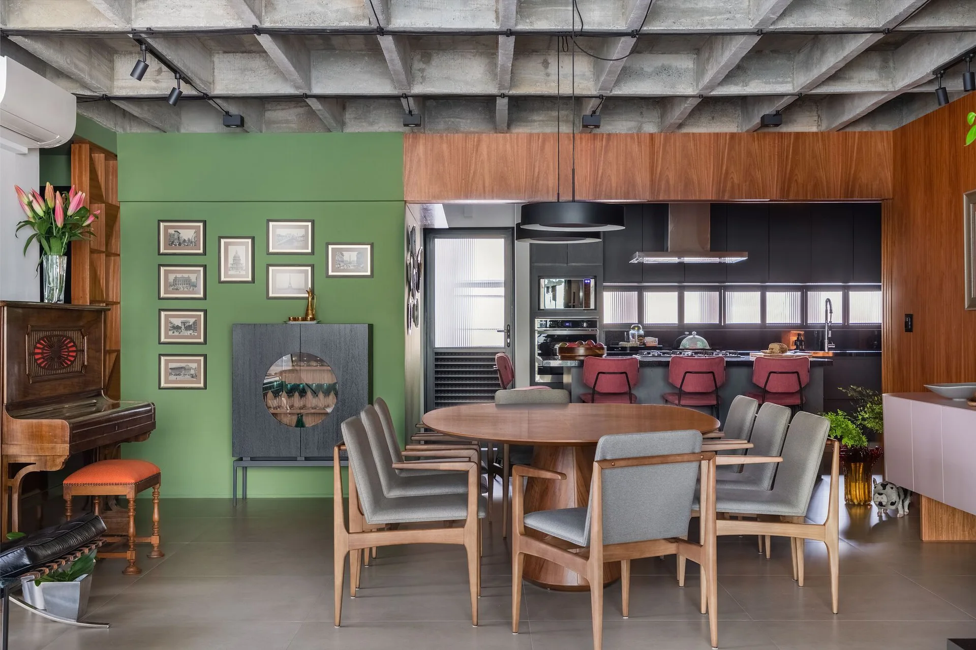 A round wooden dining table and grey chairs sit beneath an exposed concrete waffle ceiling next to a green accent wall.