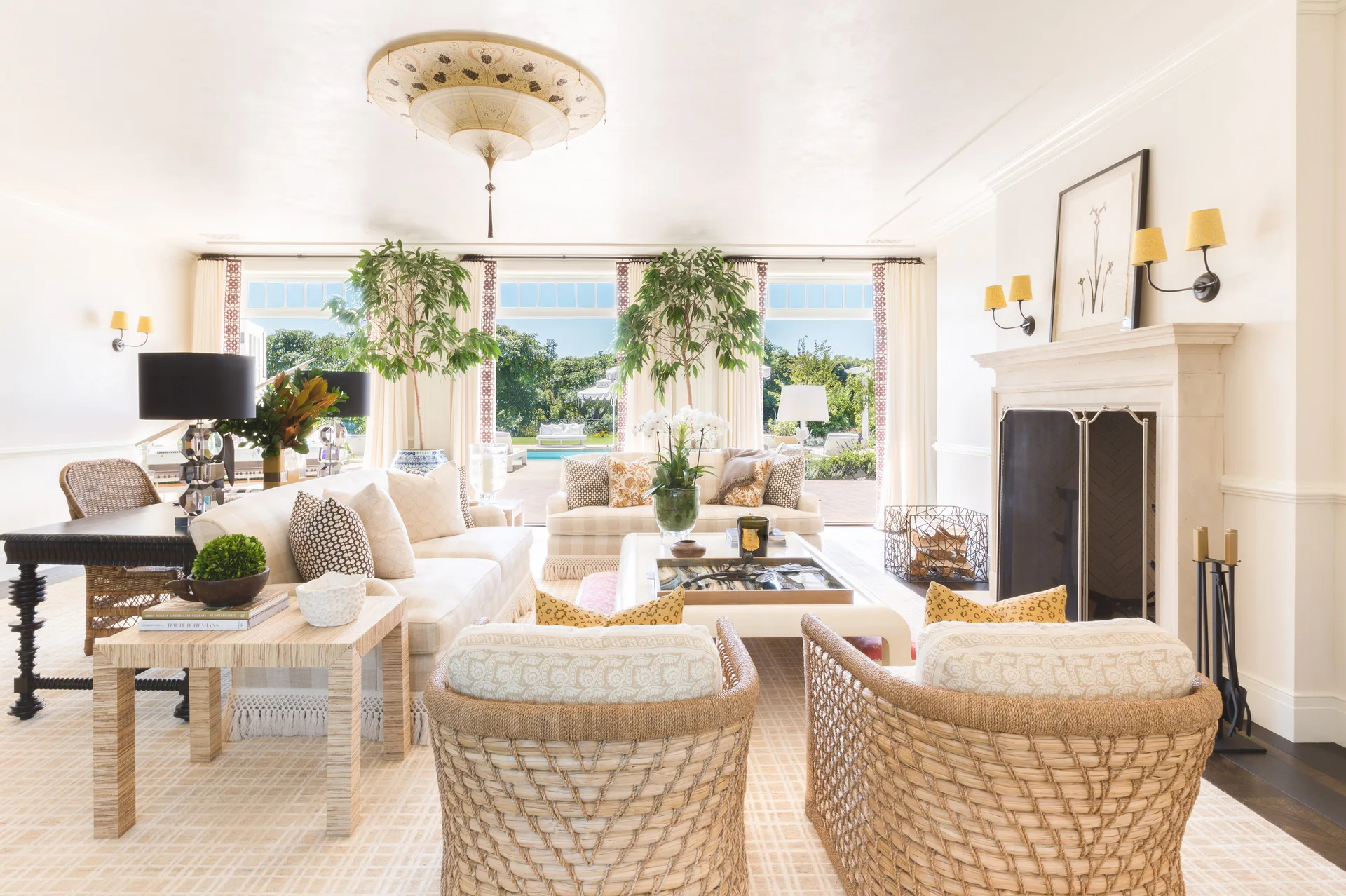 A bright coastal living room featuring two woven wicker armchairs, a white sofa, a stone fireplace, and tall potted trees framing large back windows.