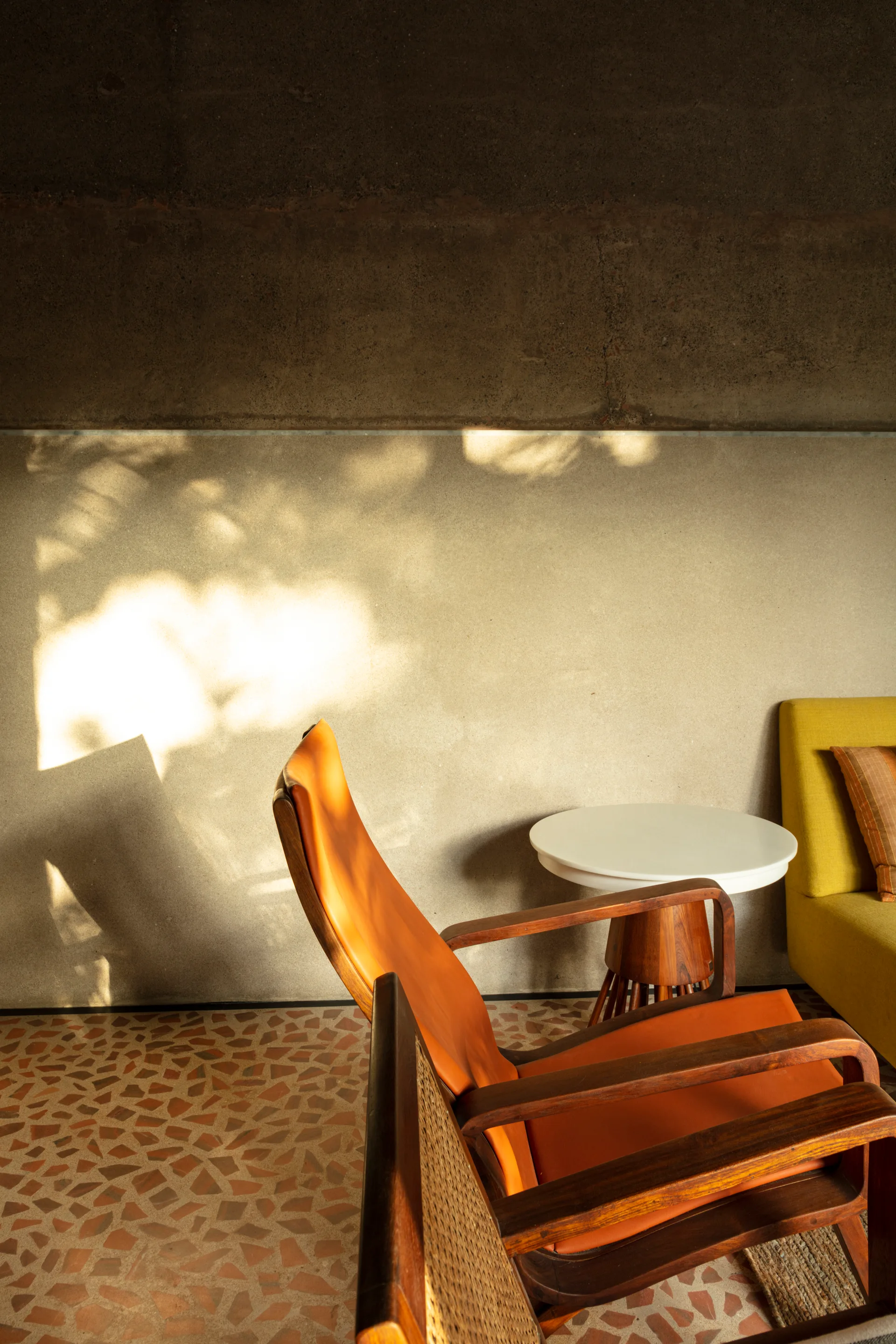An orange leather armchair with a wood frame and cane backing sits on a patterned terrazzo floor in front of a raw concrete wall with cast shadows.