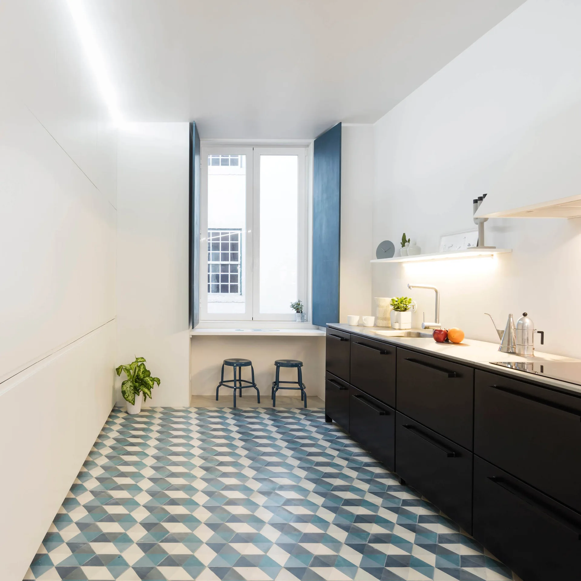 A galley kitchen featuring matte black lower cabinets, white walls, blue geometric tiled flooring, and a window with deep blue shutters.