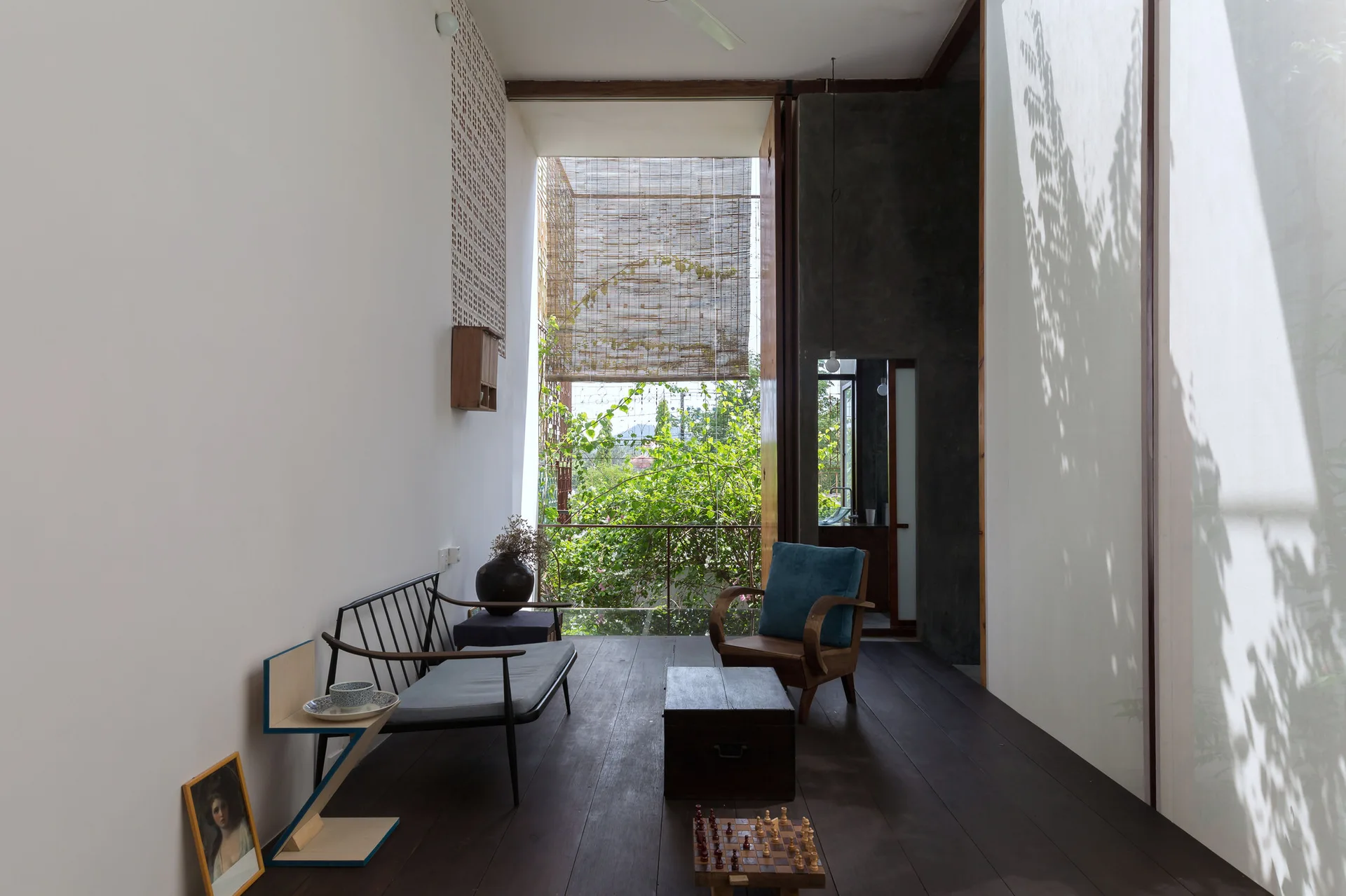 A minimalist semi-outdoor living room with a black frame sofa, wooden armchair, and low coffee table on dark wood flooring under a bamboo blind.