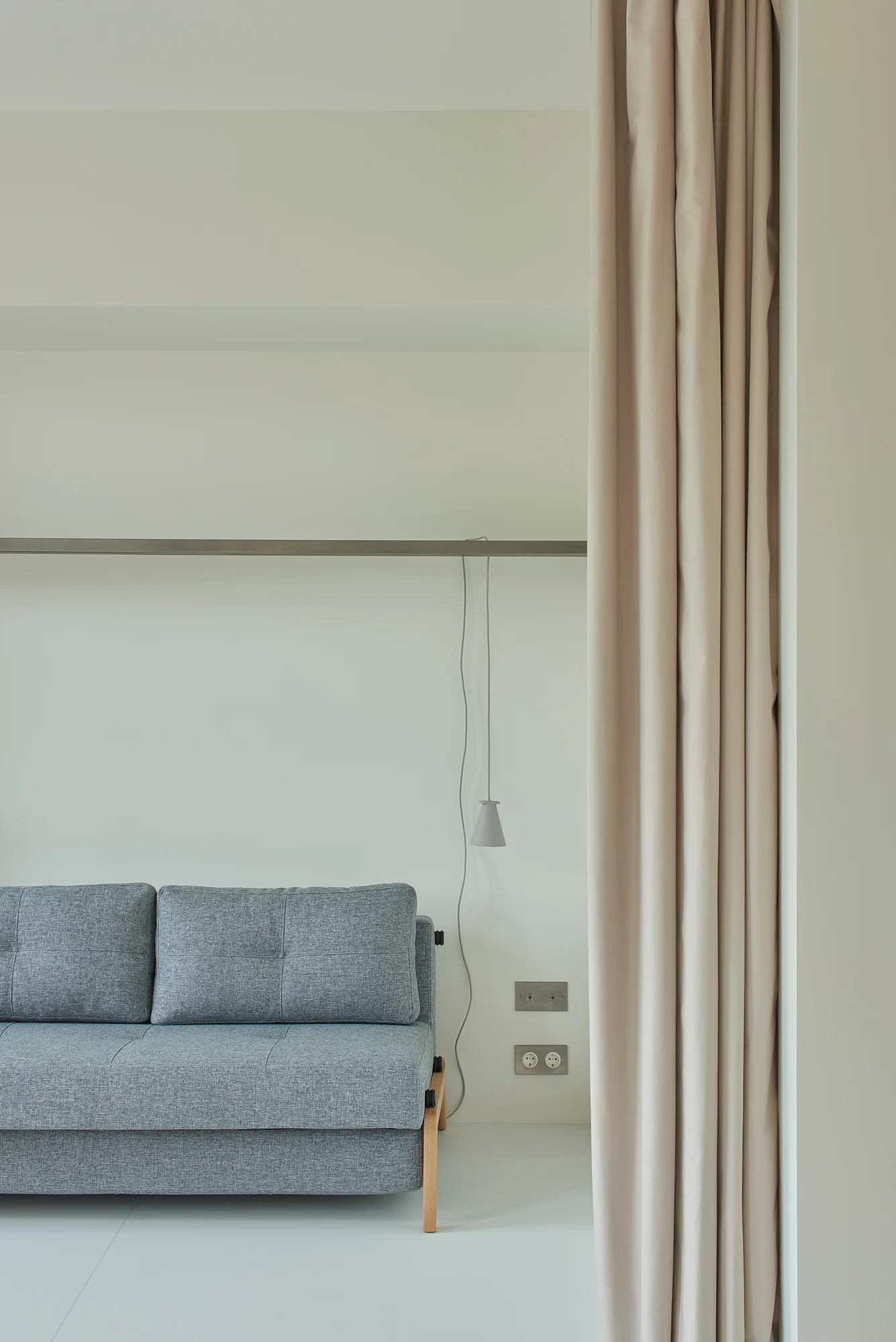 A grey upholstered sofa against a plain white wall next to a tall, draped beige curtain and a hanging pendant lamp.
