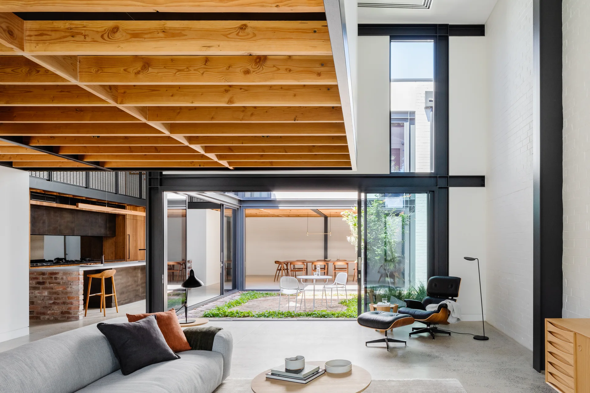 A modern living room features a gray sofa, a black leather lounge chair, and glass doors opening onto an interior courtyard under exposed wood joists.